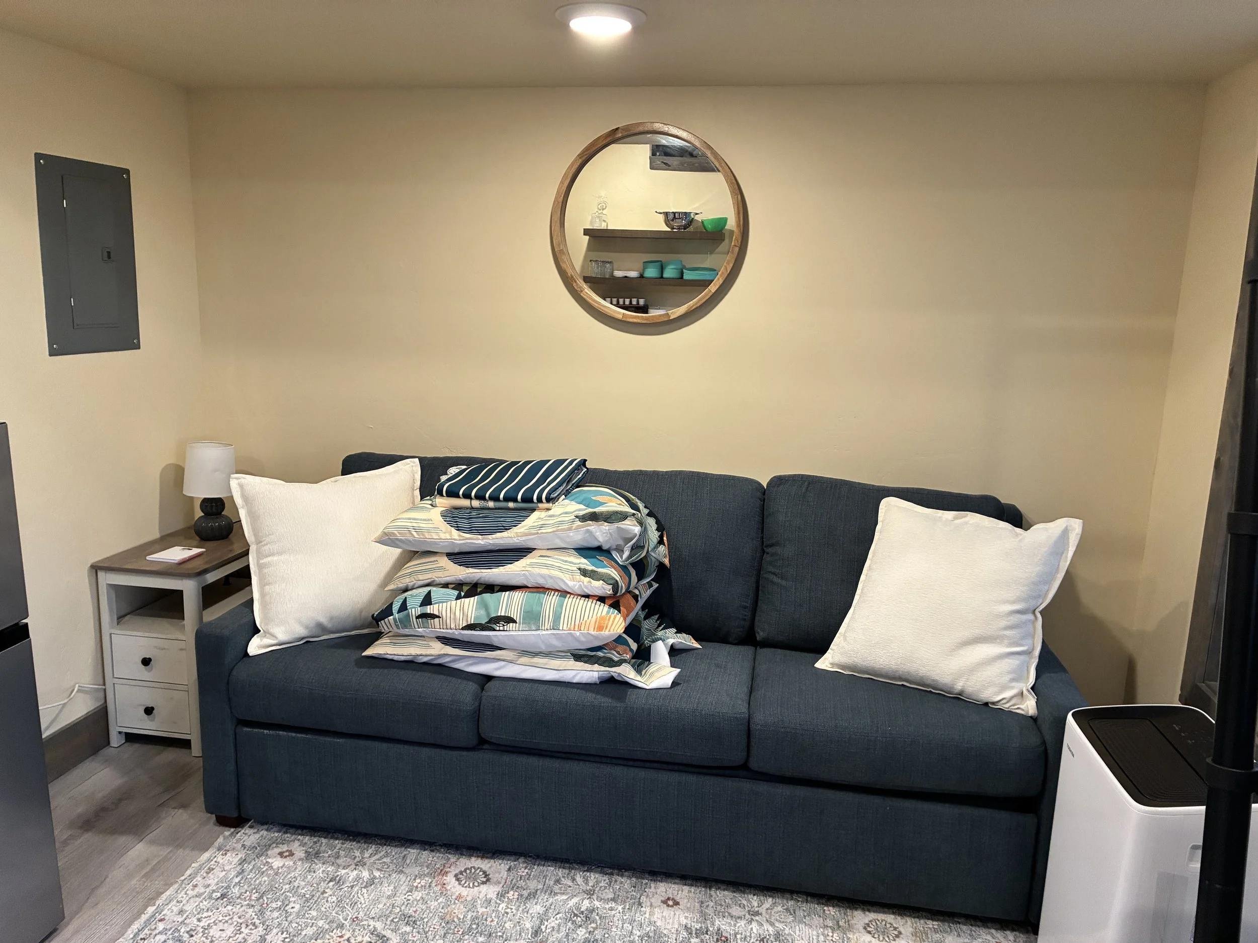 Living room with a dark blue sofa, white pillows, and a pile of pillows with colorful patterns on top. There is a small white side table with a black lamp and a book. A patterned rug covers part of the wooden floor. On the wall, there is a round wooden-framed mirror and an open shelf with bowls and cups. A white air purifier is visible at the bottom right corner.