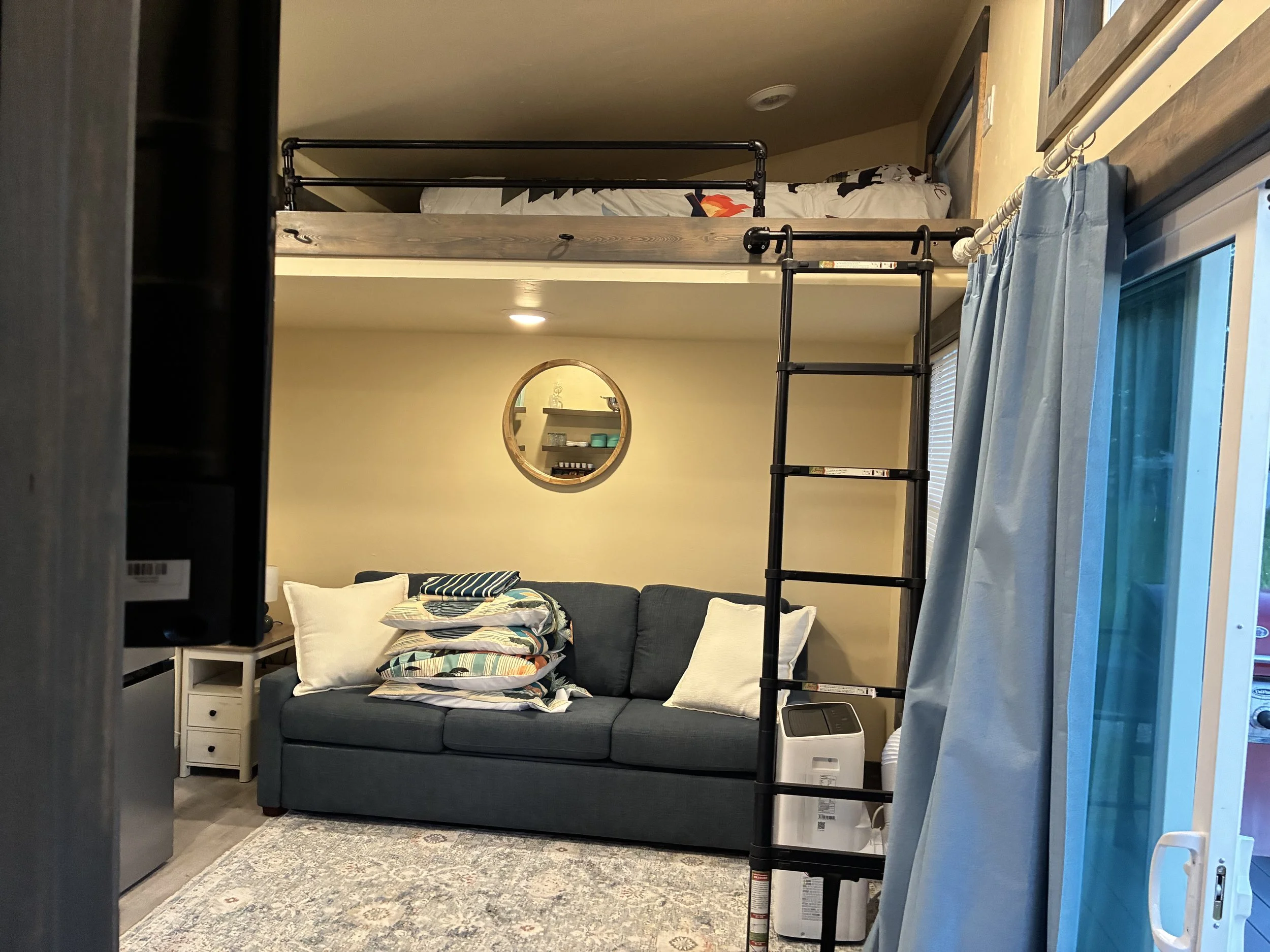 Living room with a couch stacked with pillows, a white side table, and a window with blue curtains. There is a loft bed above the living room area, accessed by a black ladder. A small mirror is on the back wall. The room has beige walls and a gray patterned rug.