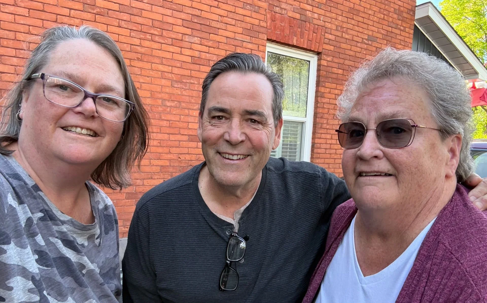 Three smiling adults posing for a photo outdoors in front of a red brick house with green trees in the background.