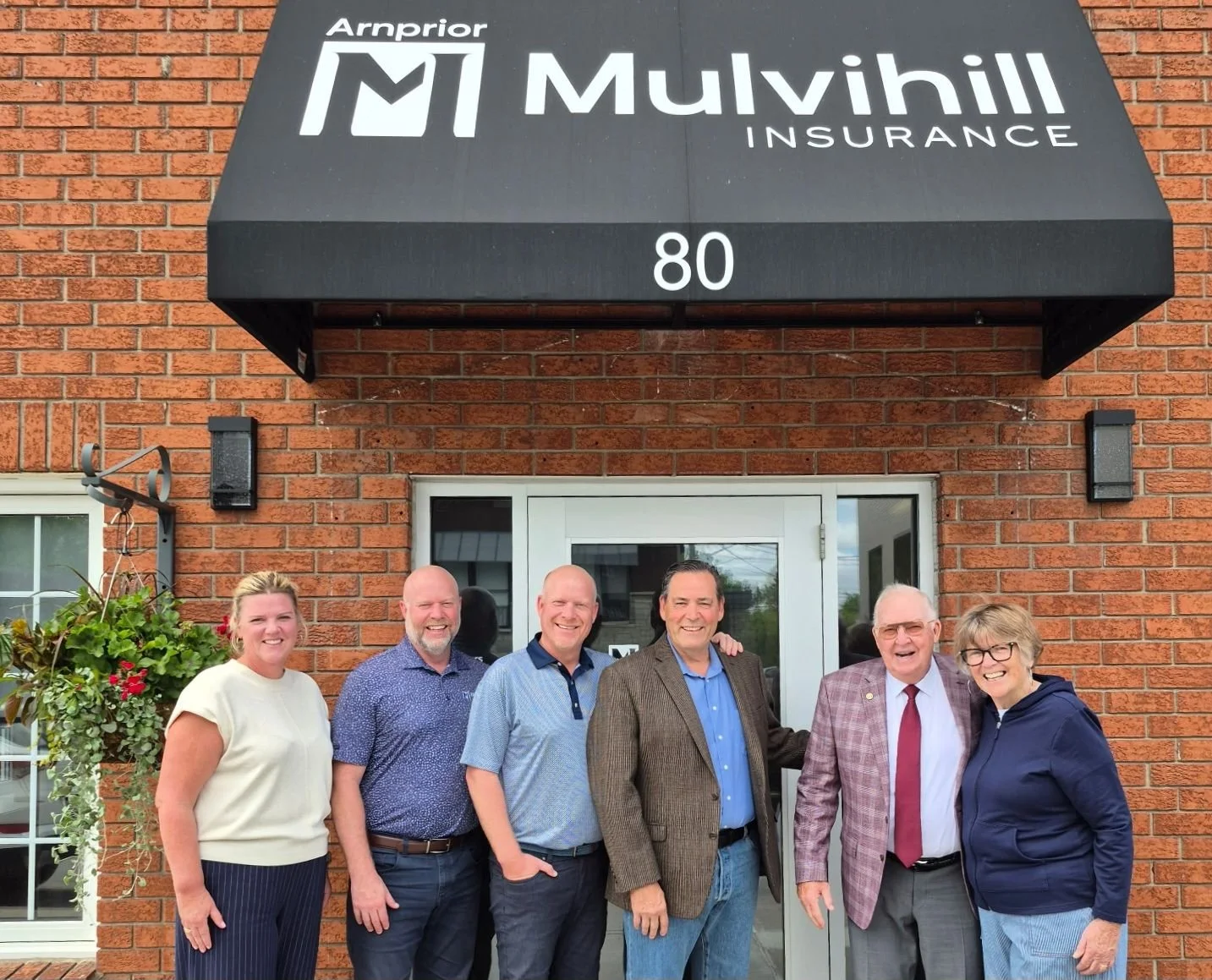Group of six people standing in front of a brick building with an awning that reads 'Arnprior Mulvihill Insurance' and the number 80. They are smiling and posing for the photo.