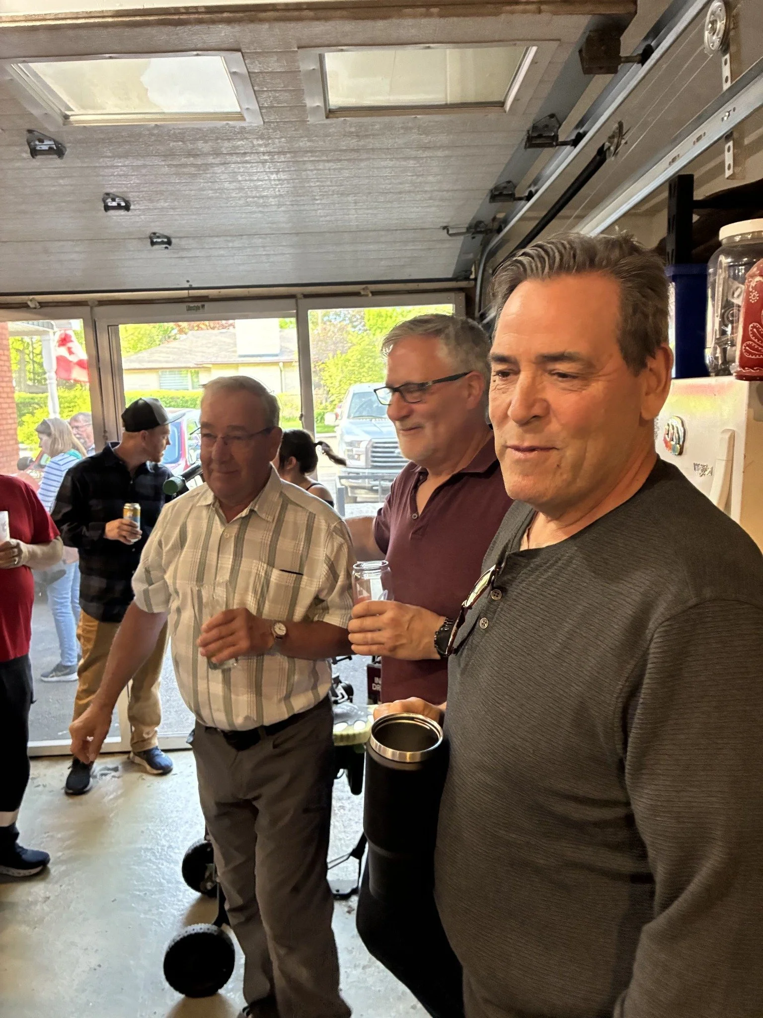 A group of older men standing inside a house with large windows, holding drinks, in a social gathering. One man is in the foreground, and others are behind him, some engaging in conversation.