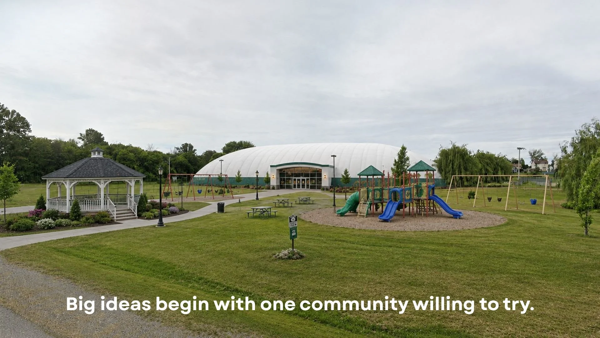 A park with a gazebo, a large indoor arena, and a children's playground with slides and swings, under a cloudy sky.