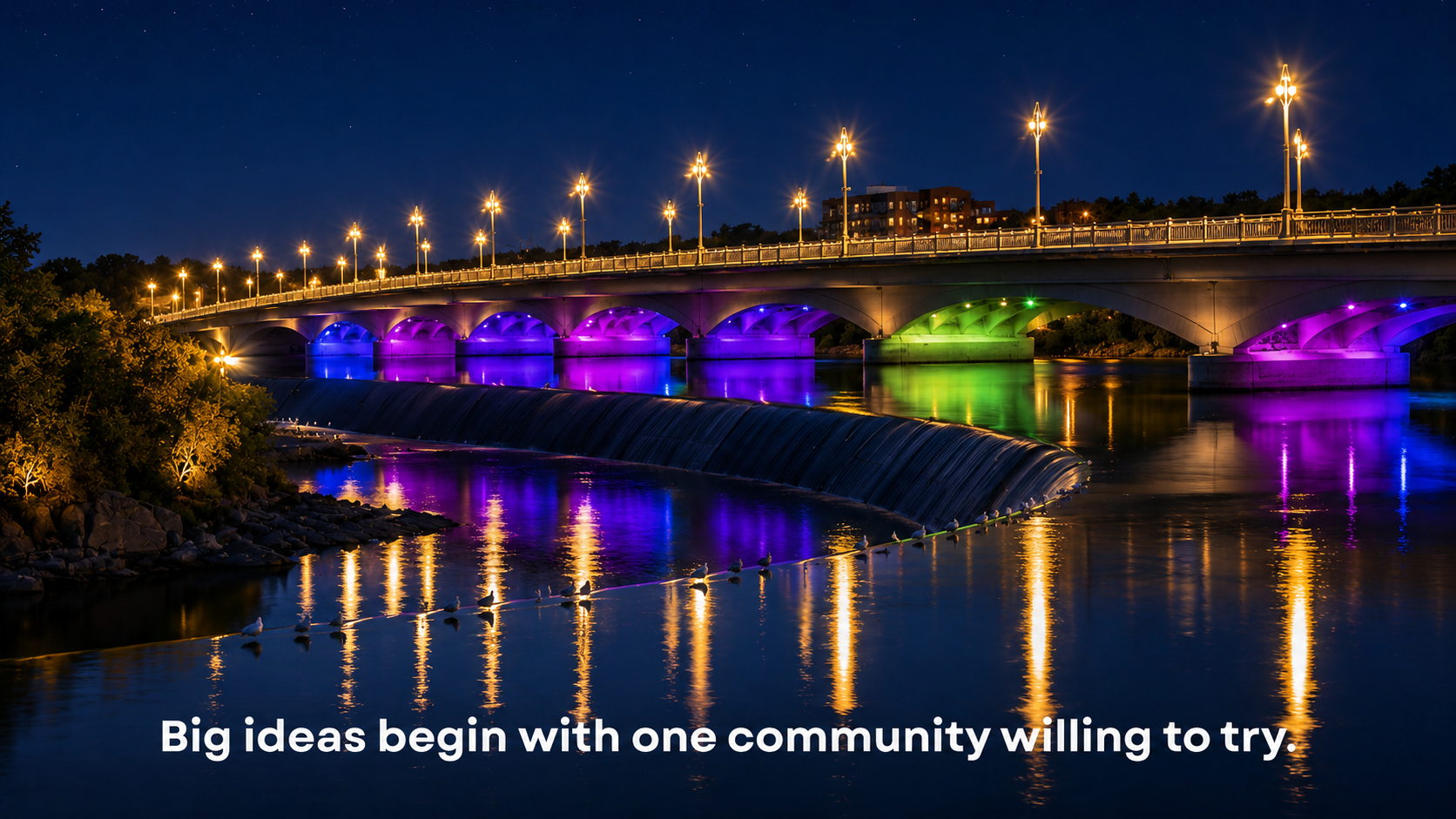 Night scene of a brightly lit bridge over a river with colorful LED lights beneath the arches, reflecting on the water, and a quote that says, 'Big ideas begin with one community willing to try.'