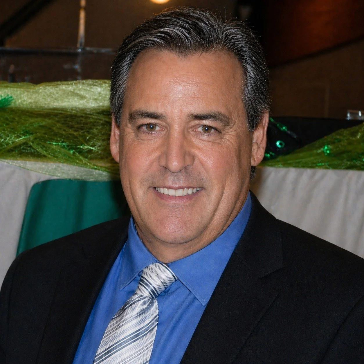 A middle-aged man in a suit smiling, with dark hair streaked with gray, in front of a decorated background at an indoor event.