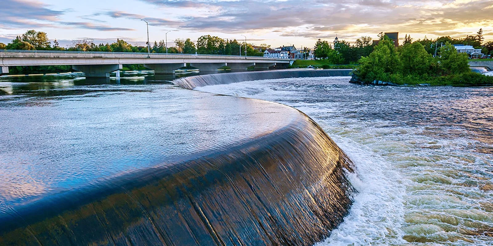 A river with a curved weir or dam, a bridge across the river, and a small island with trees, buildings, and a sunset sky in the background.