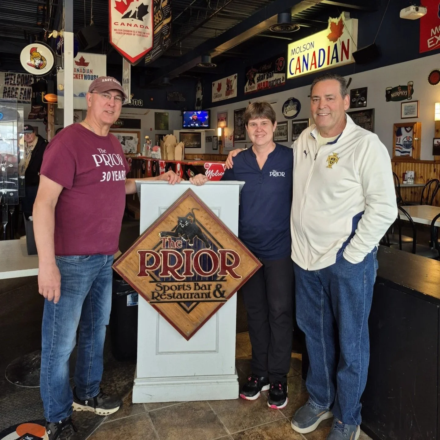 Four people smiling and standing around a sign that reads 'The PRIOR Sports Bar & Restaurant' inside a sports bar, with various signs and a TV showing sports in the background.