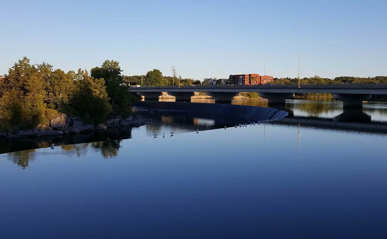 Calm river with a small waterfall, trees on the left, and a bridge over the river, with buildings in the background under a clear blue sky.
