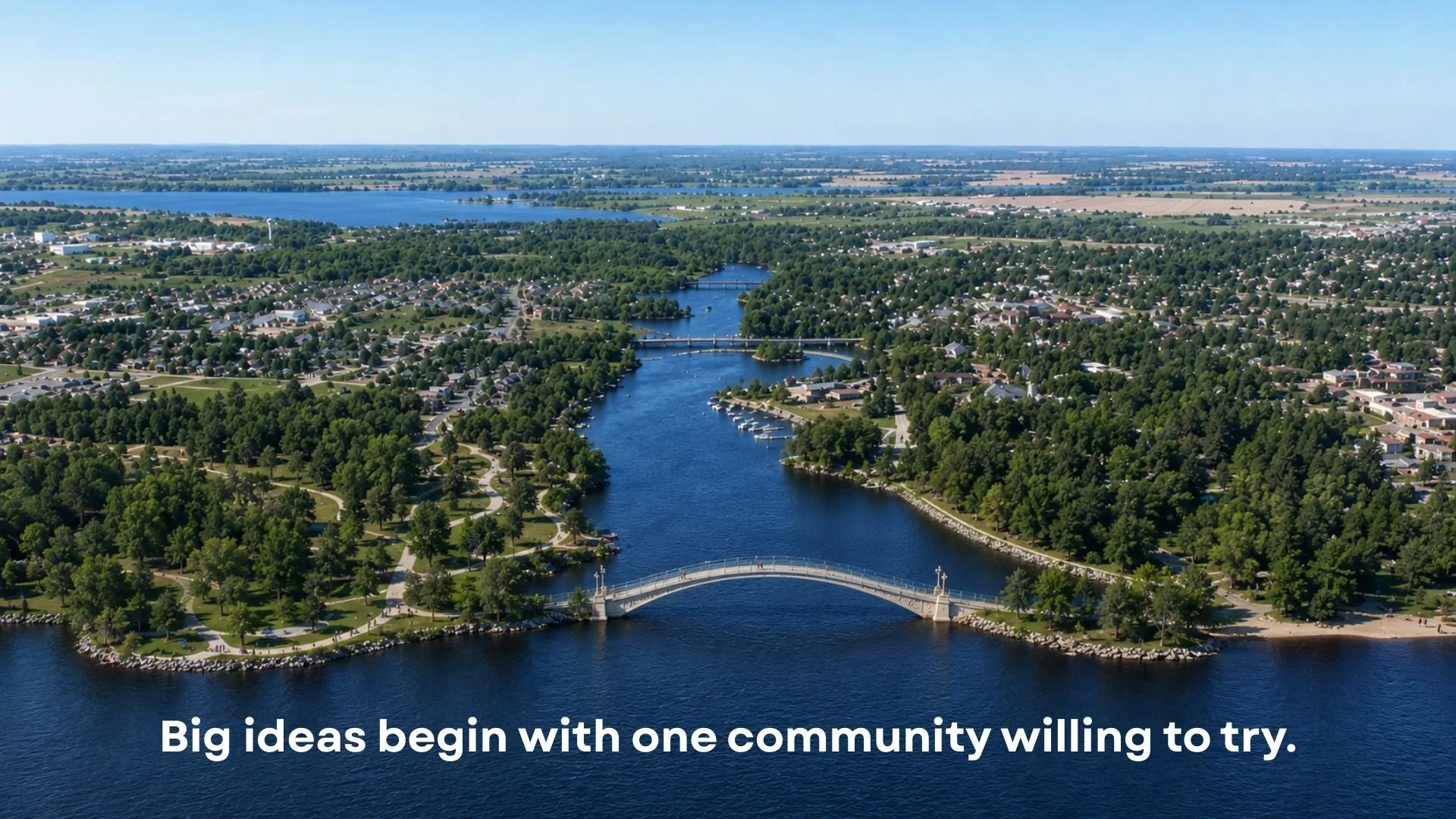 Aerial view of a small town with a winding river flowing through it, multiple bridges crossing the river, surrounded by lush greenery and residential neighborhoods, with a lake in the distance under a clear blue sky.