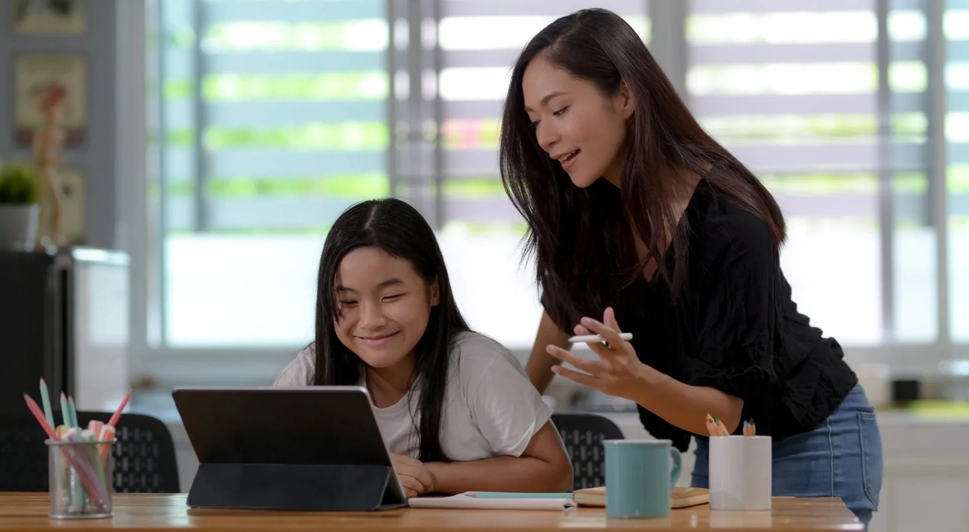 A young girl with long dark hair sitting at a desk, smiling at a tablet, while an adult woman with long dark hair and wearing a black top leans over, holding a pencil and engaging with her.