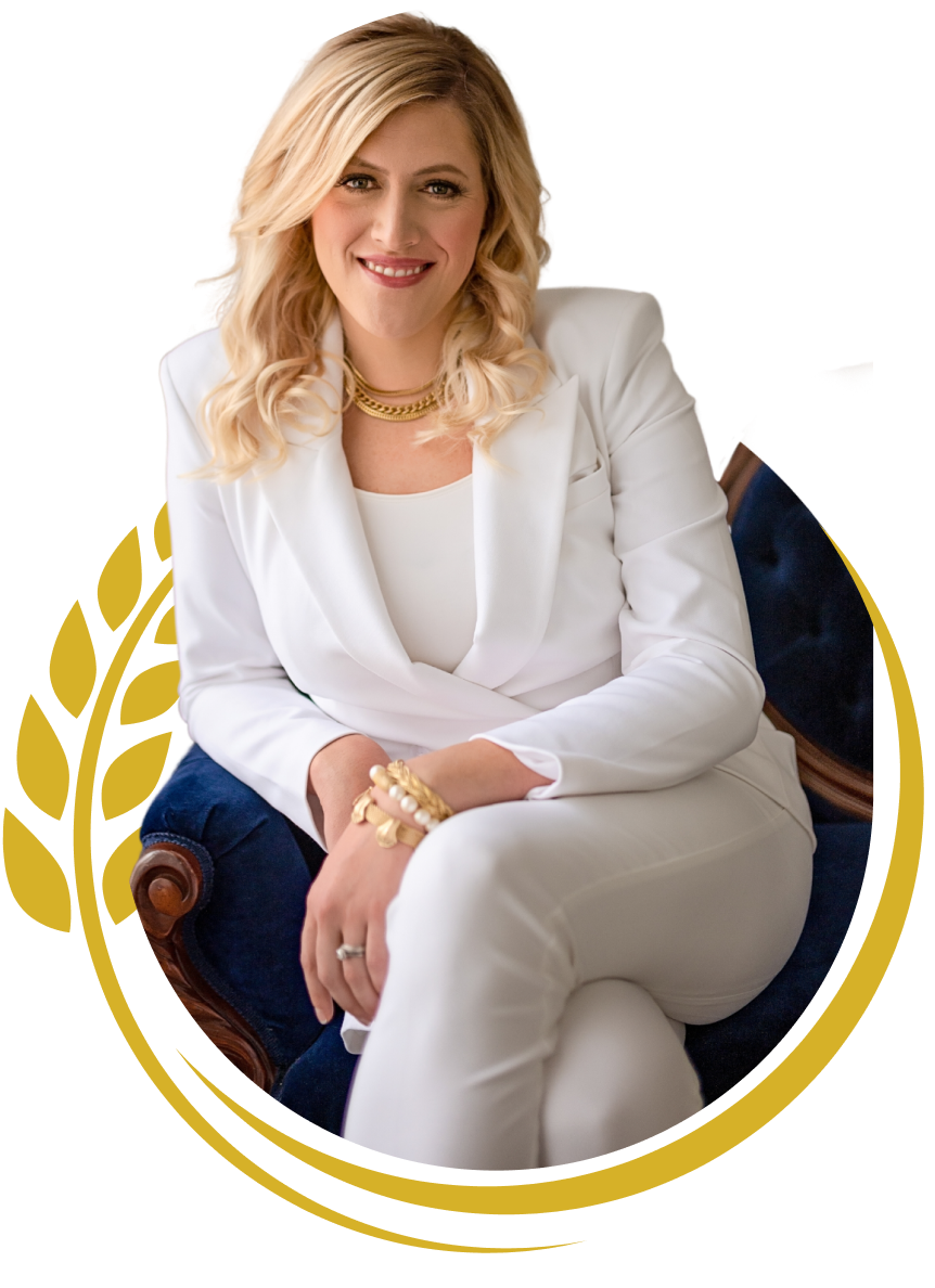 A smiling woman with blonde hair, wearing a white blazer and matching pants, sitting on a navy blue armchair with wooden accents, accessorized with gold jewelry, in a professional setting.