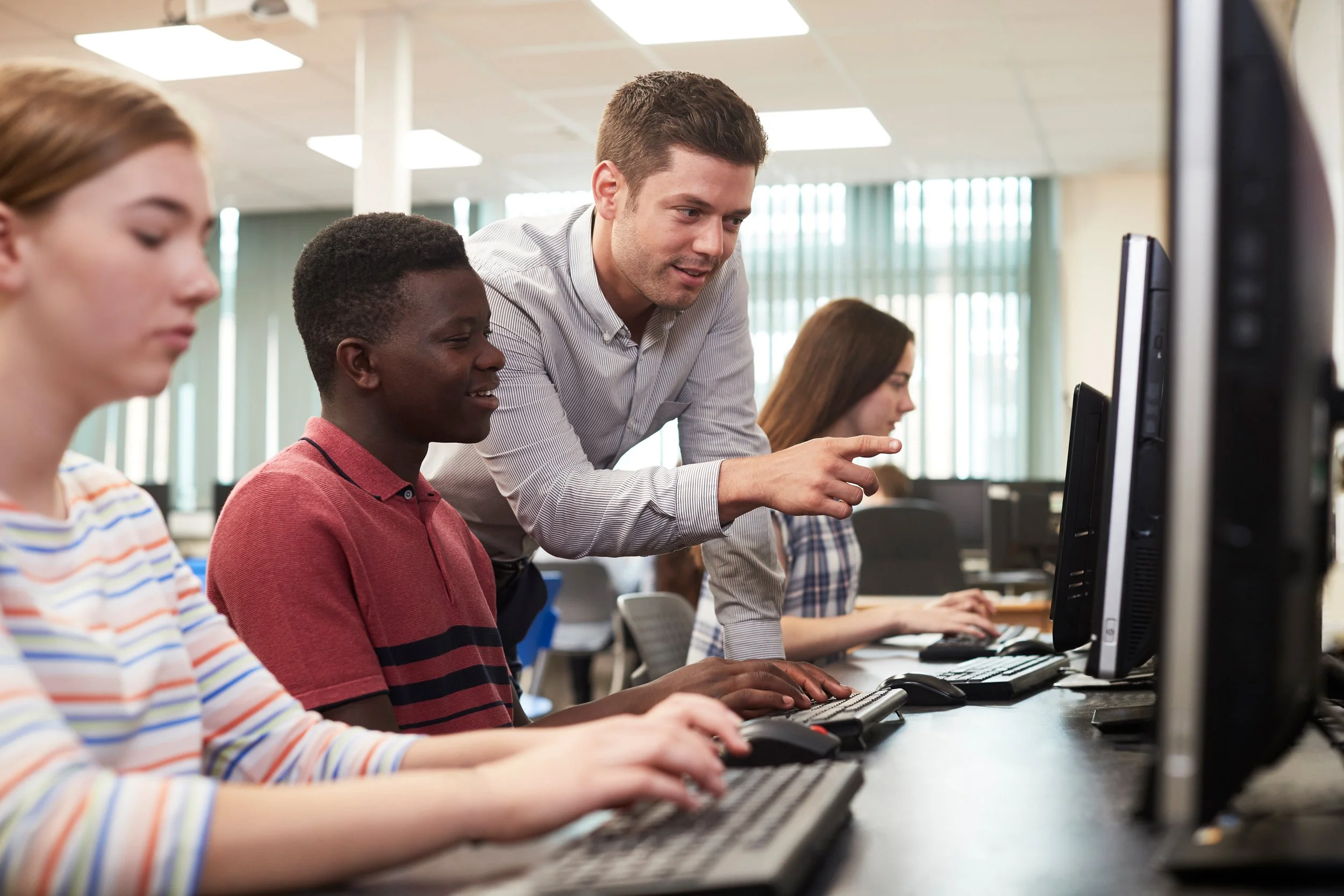 A teacher helping students in a computer lab, with students focused on their computer screens.