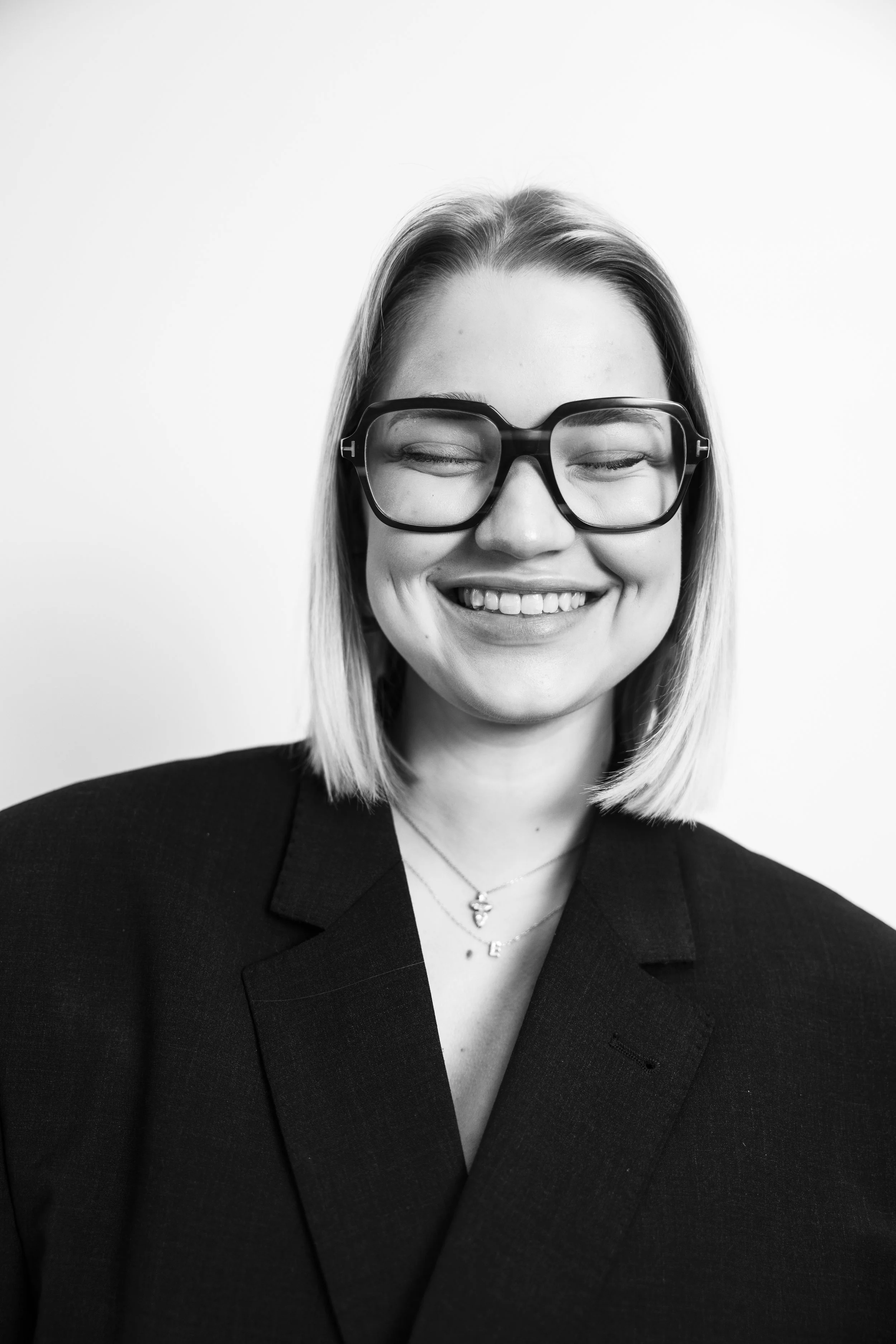 Black and white portrait of a smiling woman with straight, shoulder-length hair, wearing large glasses and a dark blazer, against a plain white background.
