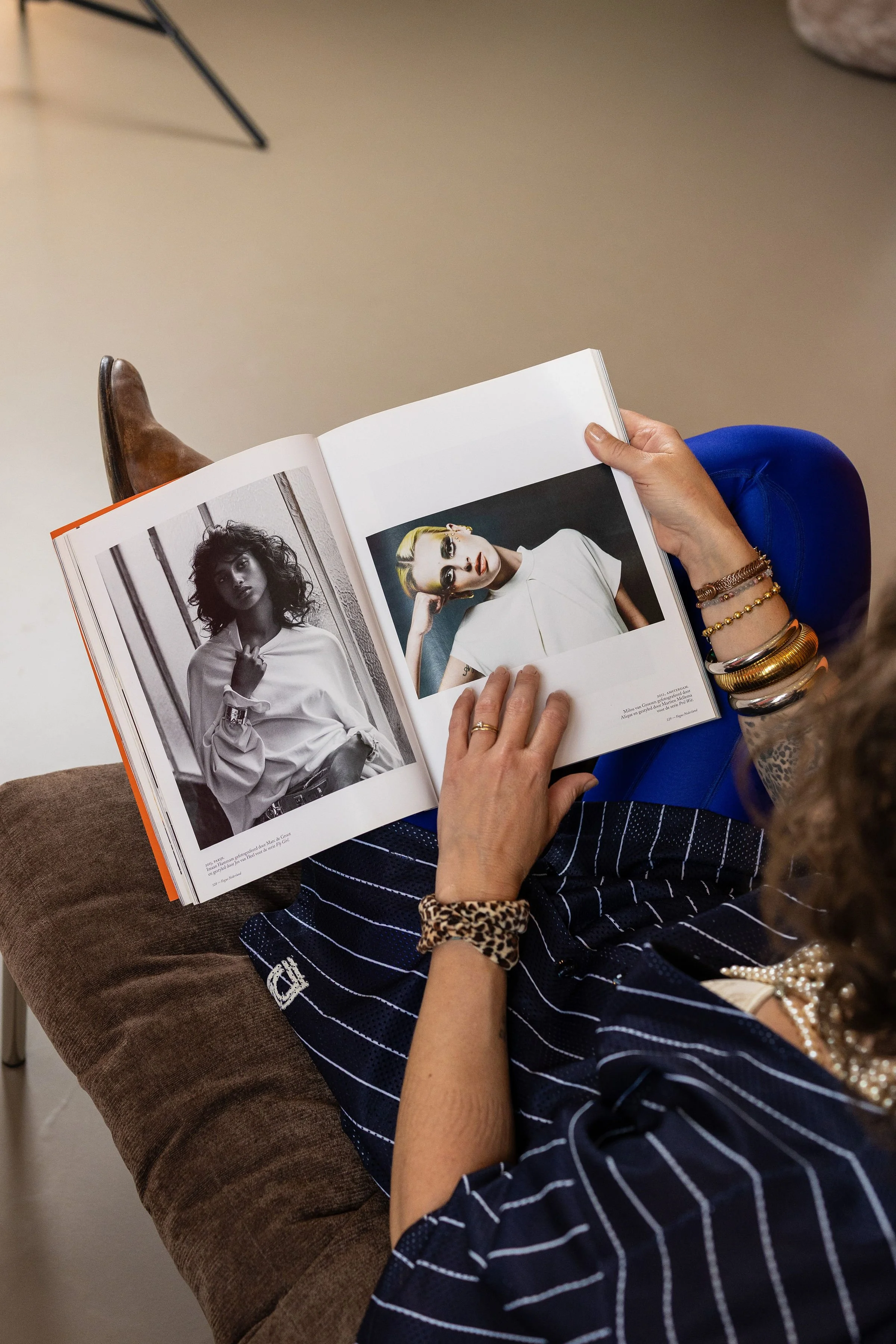 A person reading a fashion magazine with photos of models. The person is sitting on a brown chair, wearing a blue and white striped dress, and accessorizing with multiple bracelets.