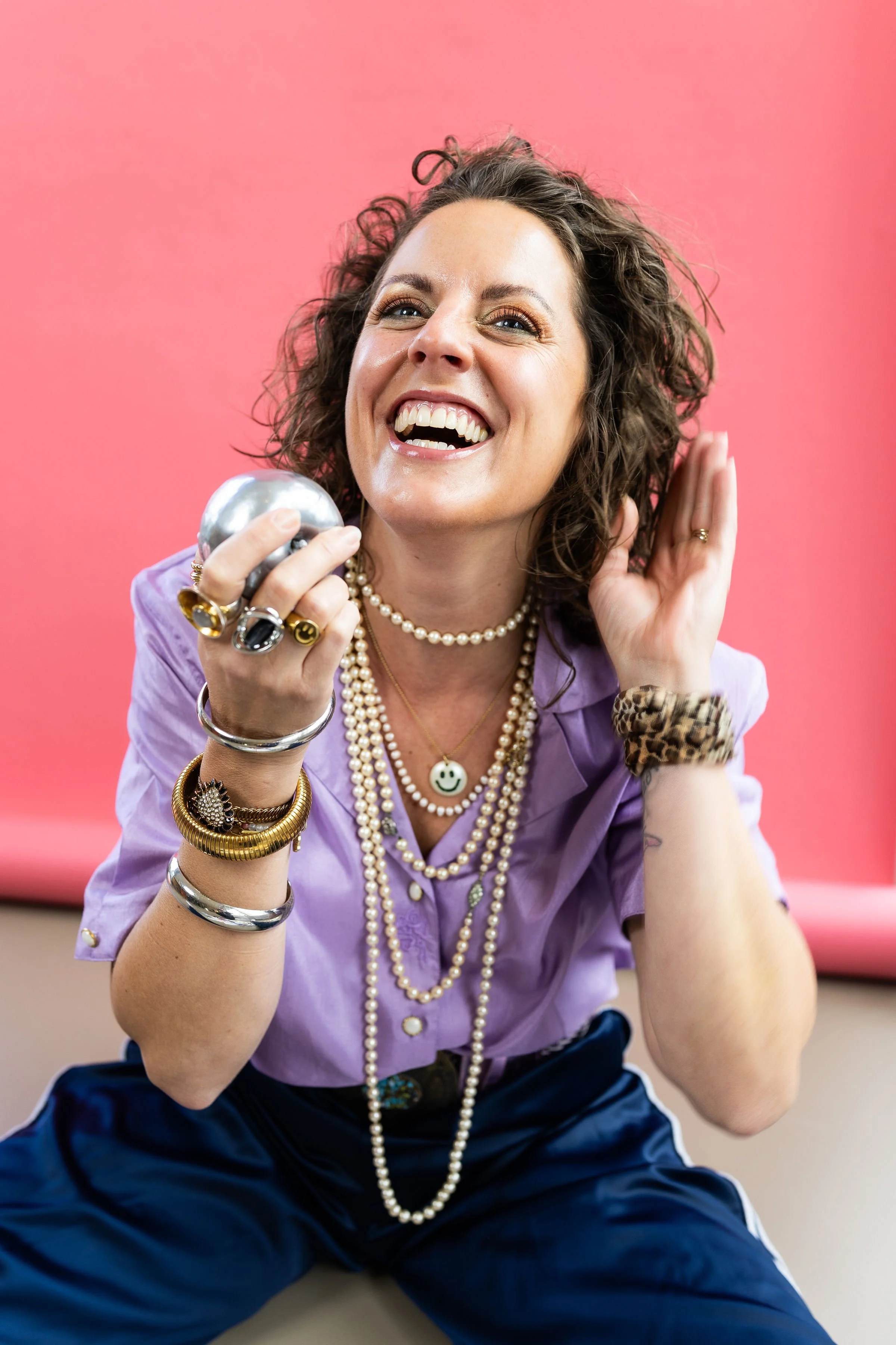 A woman with curly brown hair and bright eyes smiling broadly, holding a shiny silver ball in her hand, wearing multiple pearl necklaces, several rings, bracelets, and a leopard print scrunchie, against a pink background.