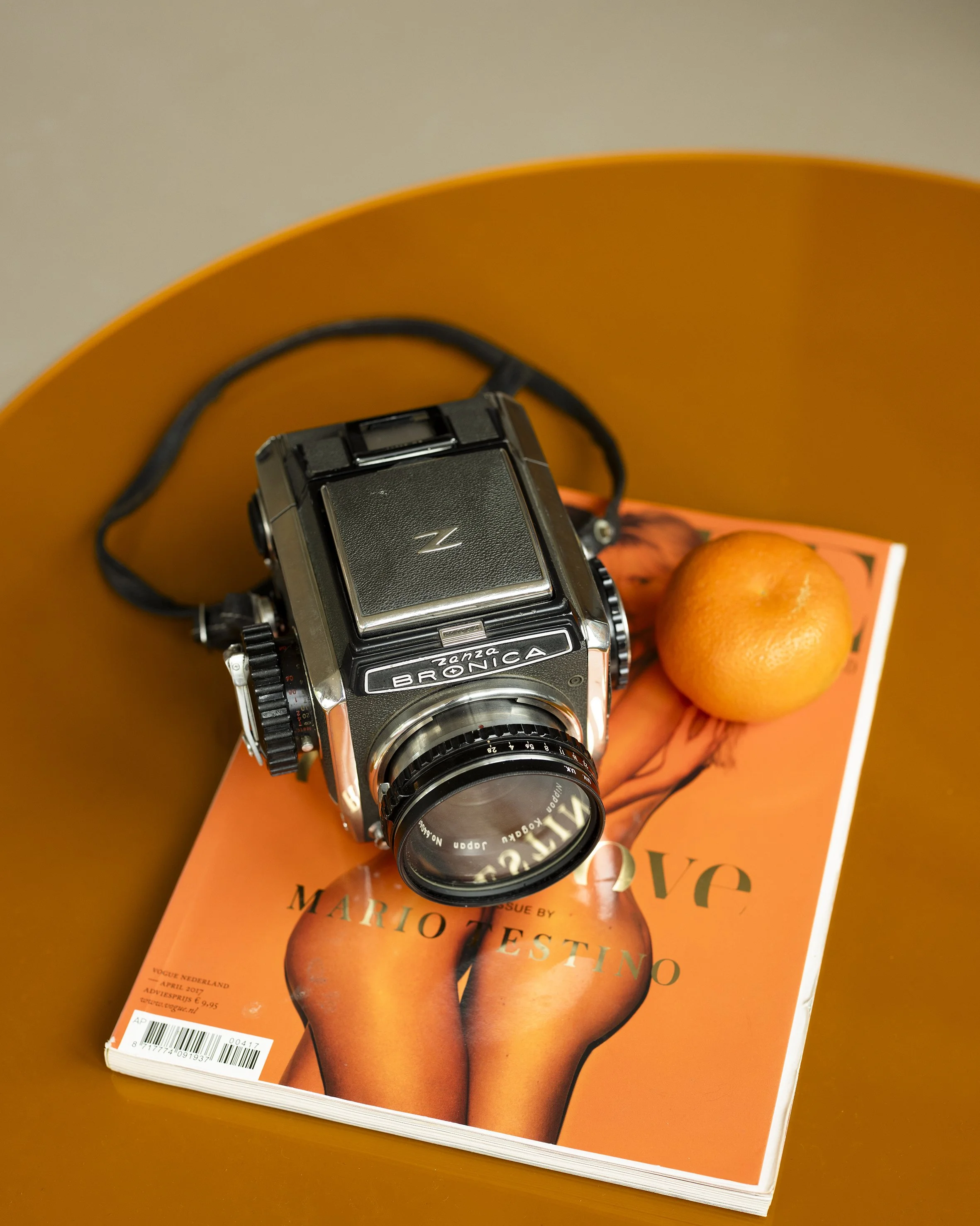 Close-up of a vintage Motorola ZandZonica pocket camera resting on a Marie Claire magazine with a model wearing orange tights and orange shoes on the cover. The scene is set on a brown surface.
