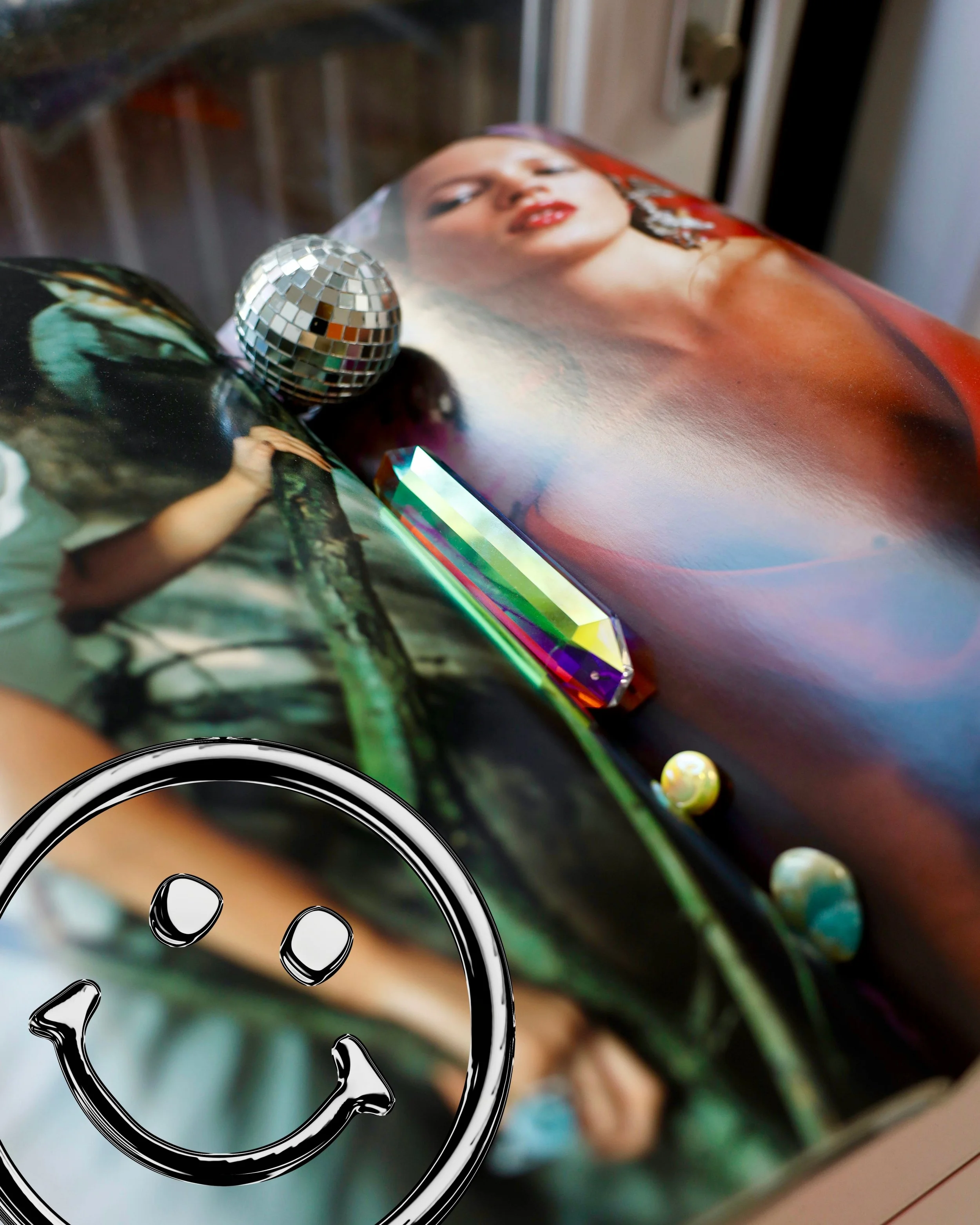 Close-up image of two magazines with a disco ball and a colorful crystal pen placed on top. The magazine on top has a woman’s face with red lipstick, and the background features a portrait with a skin tone that is slightly out of focus.