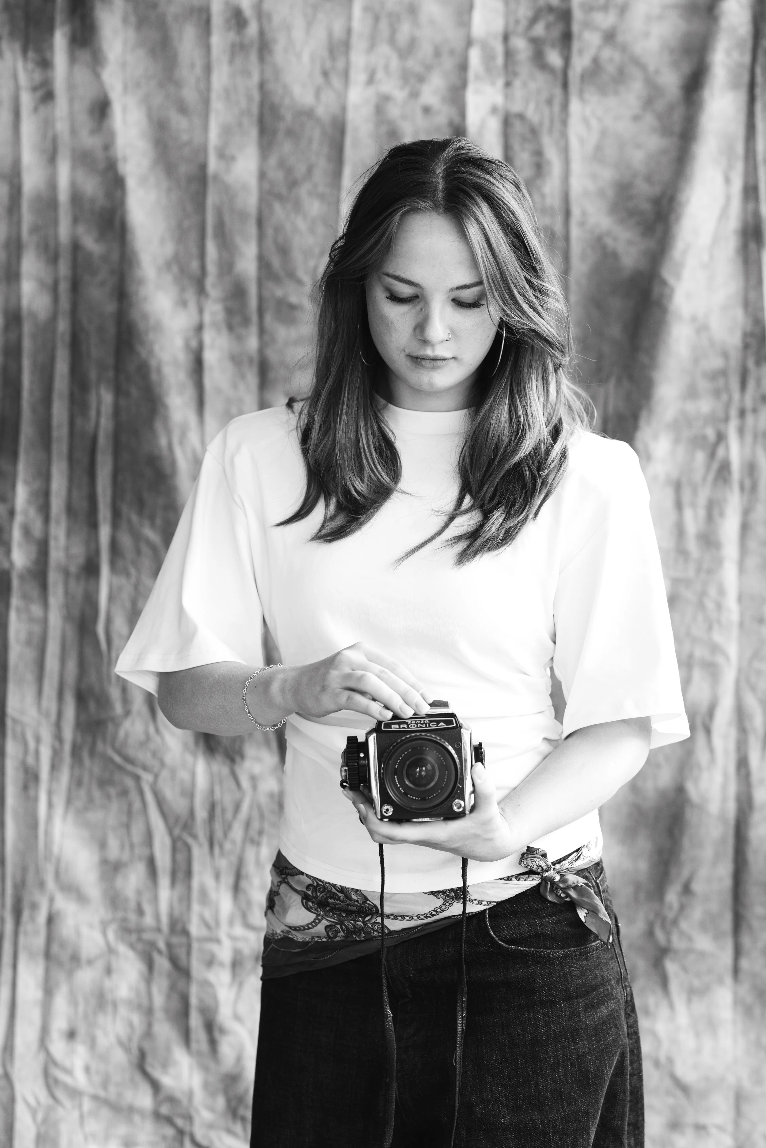 A young woman with long hair, wearing a white top and dark pants, holding an old fashioned camera in front of a wooden background.