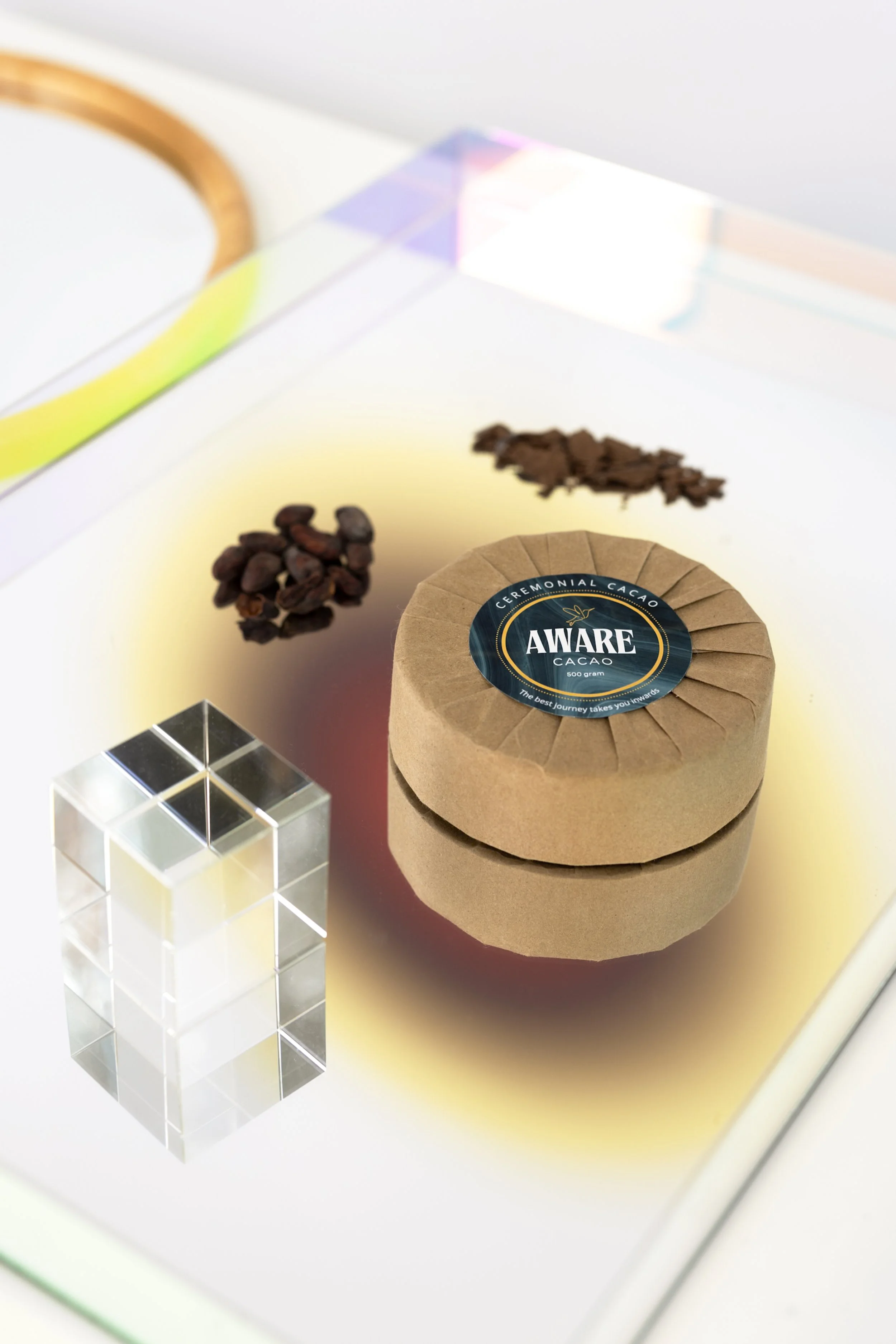 Close-up of a round package of Aware Cacao ceremonial cacao, a small pile of cacao nibs, and a clear cube, all on a glass surface with a yellow background.