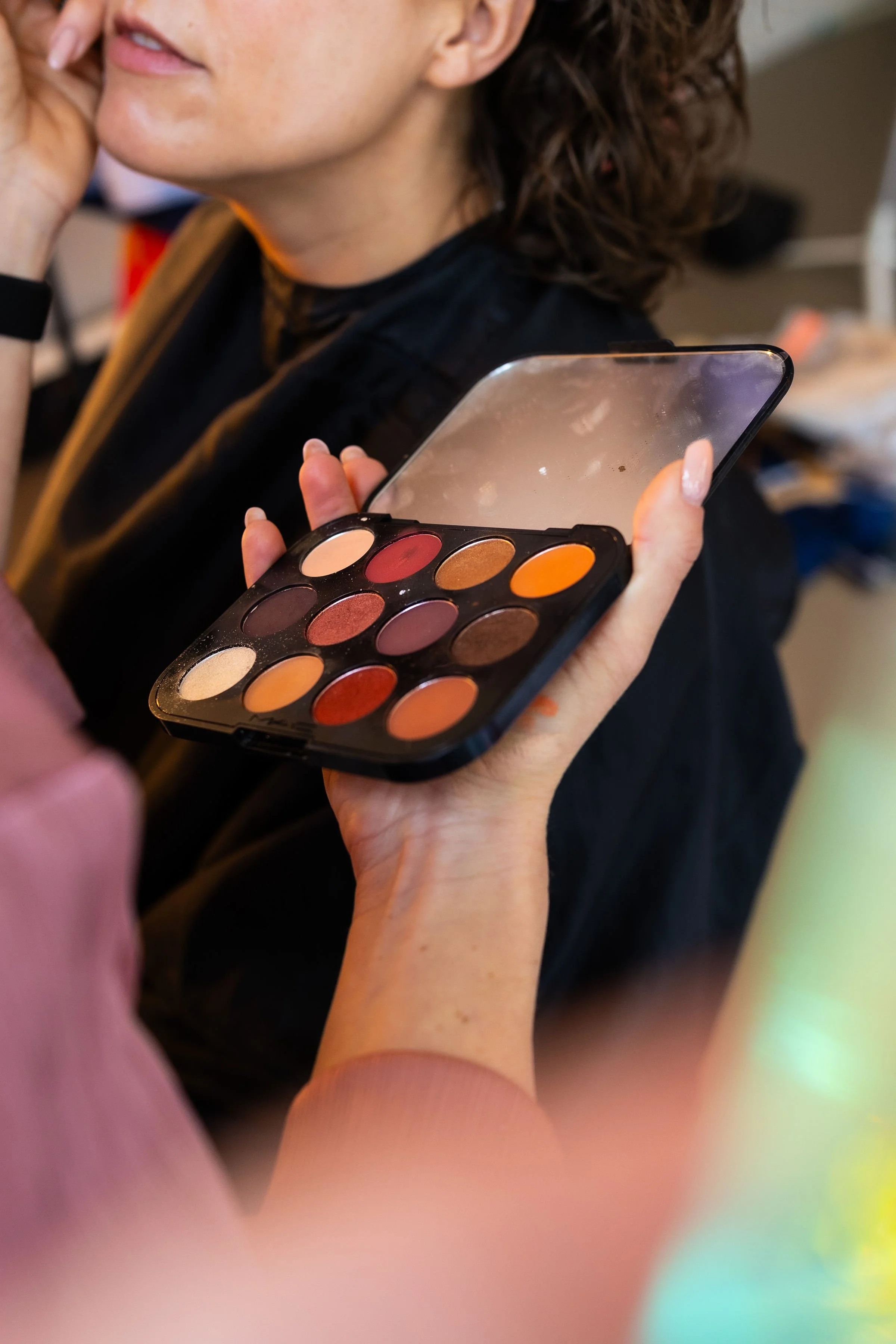 A woman with curly hair applies eyeshadow using a palette with multiple colors, with another person holding the palette.
