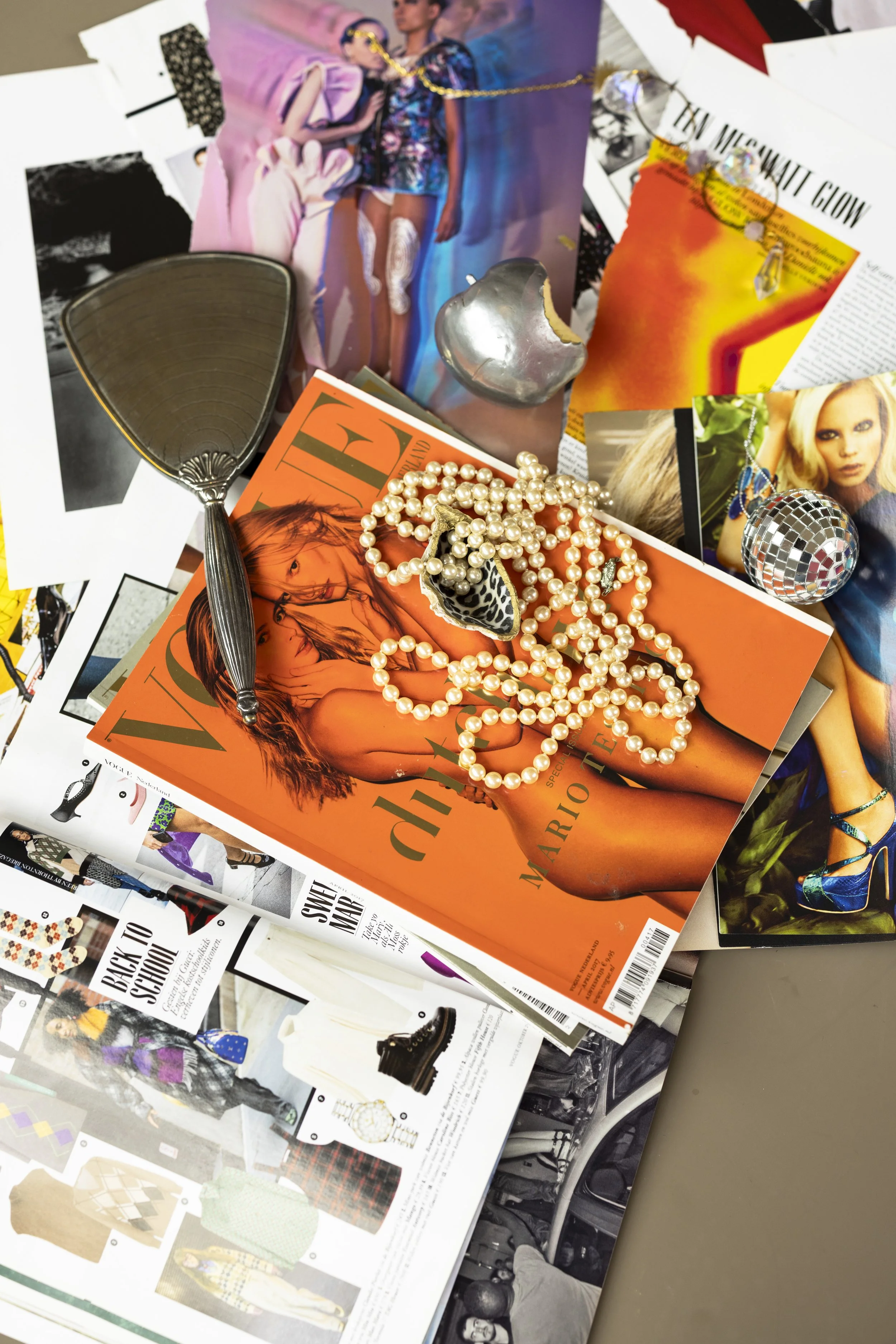 A cluttered table with various fashion magazines, jewelry, a mirror, and a partially eaten silver snack.