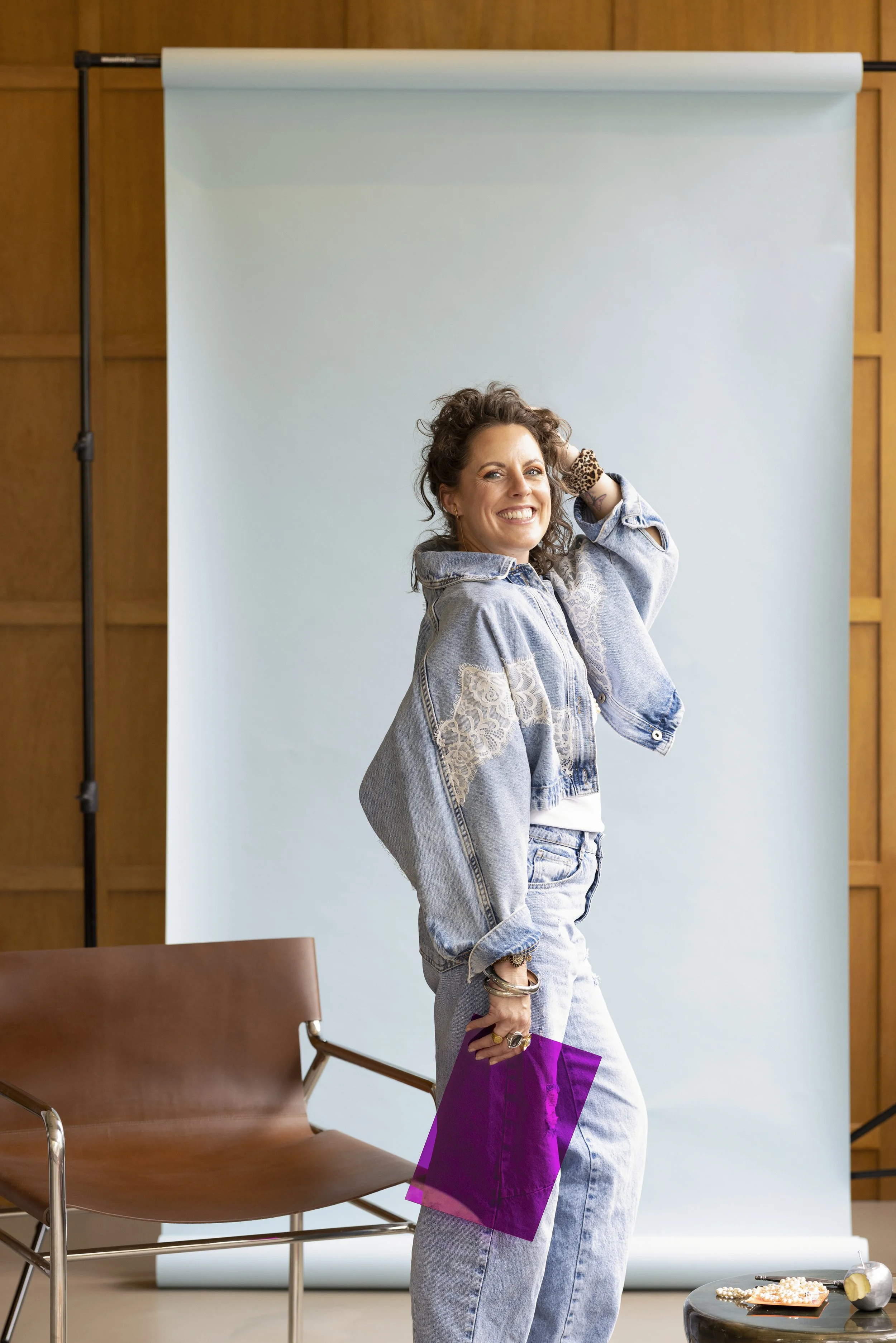 Woman smiling and posing in front of a light blue backdrop, wearing a denim jacket and light blue jeans, holding a purple and pink shopping bag, with chairs and a small table with jewelry in front of her.
