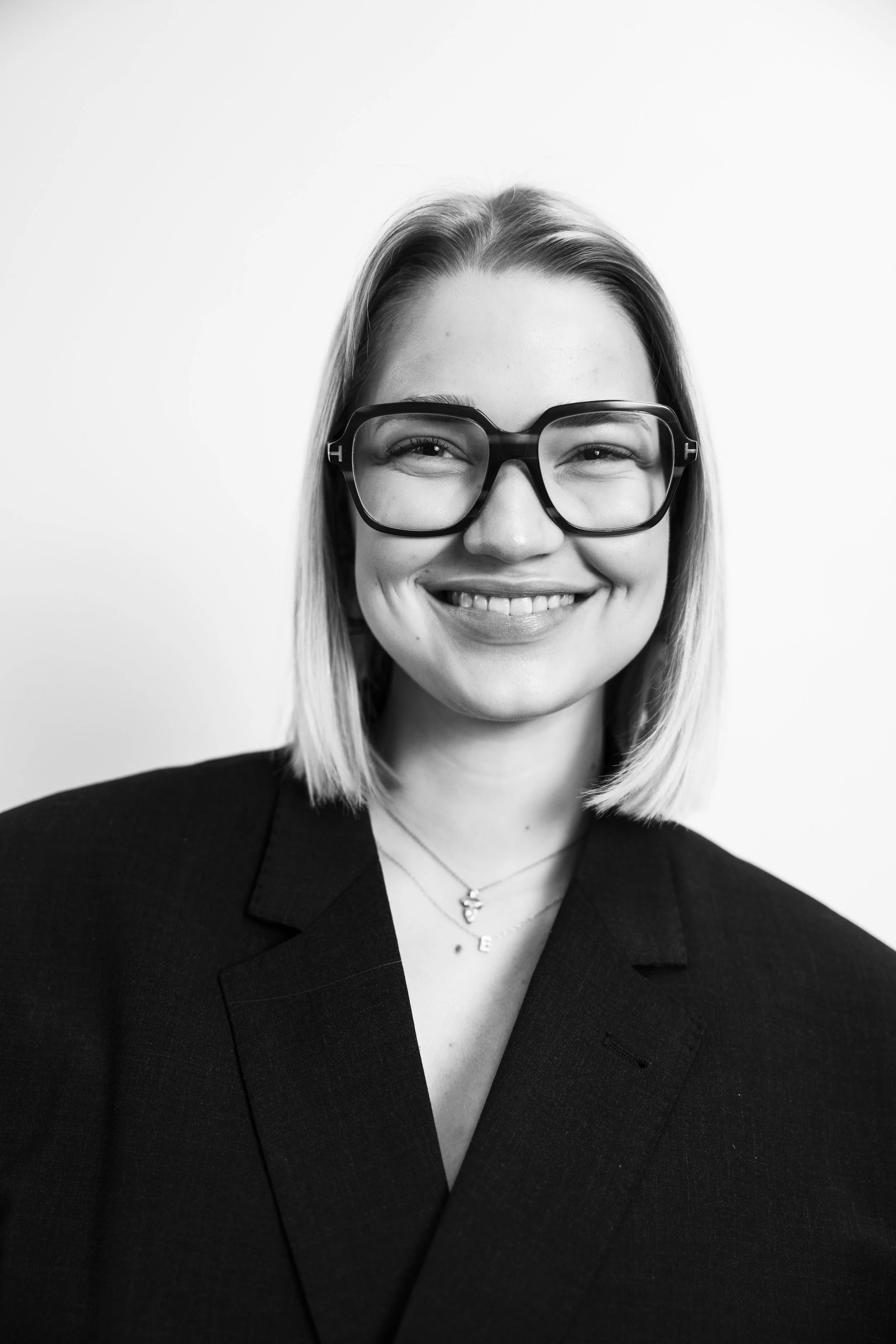 Black and white portrait of a woman with shoulder-length hair wearing large glasses, a blazer, and a heart necklace, smiling.