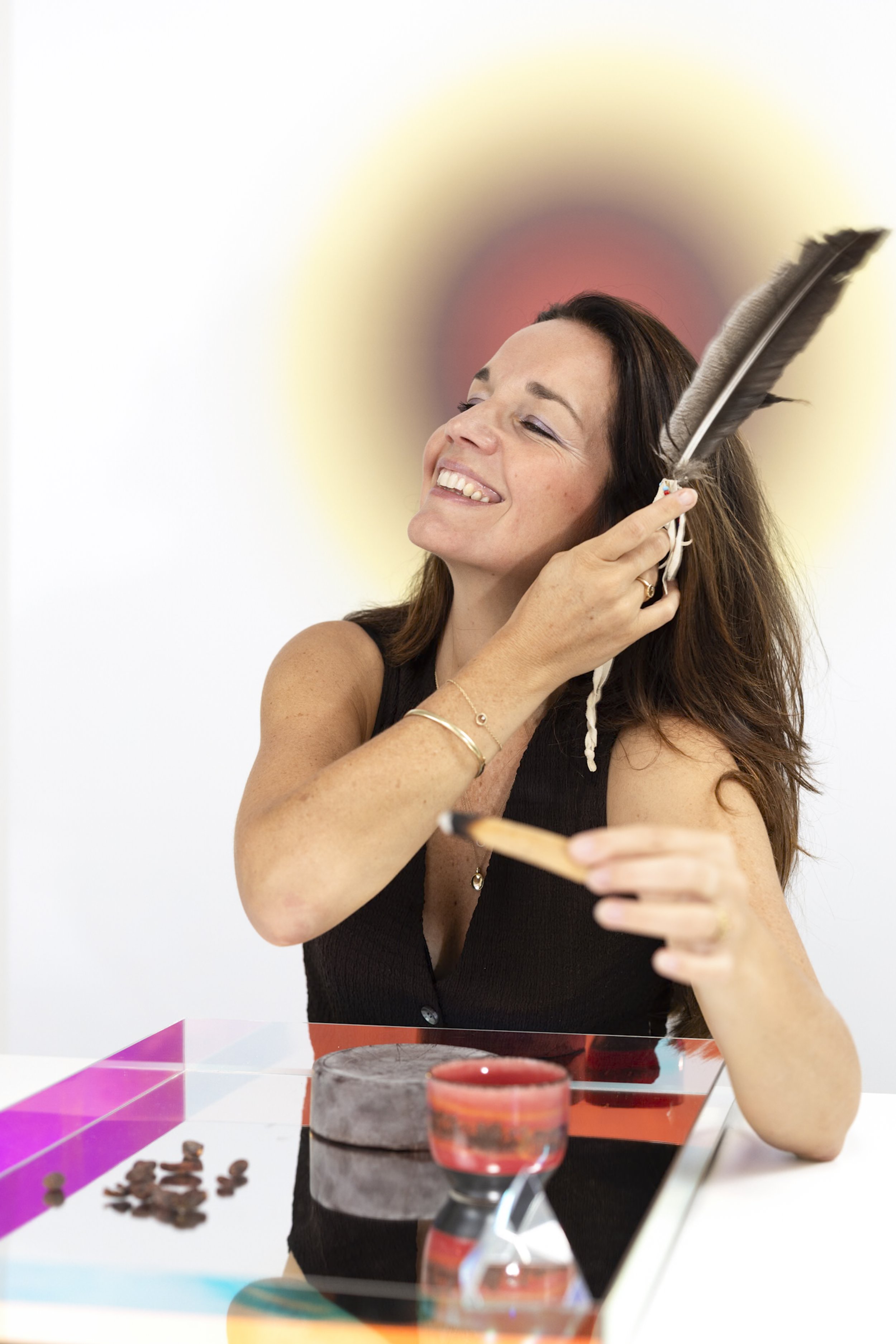 Woman smiling and smelling a chocolate cake while sitting at a table with chocolate treats and a chocolate cake in front of her, in a bright room.