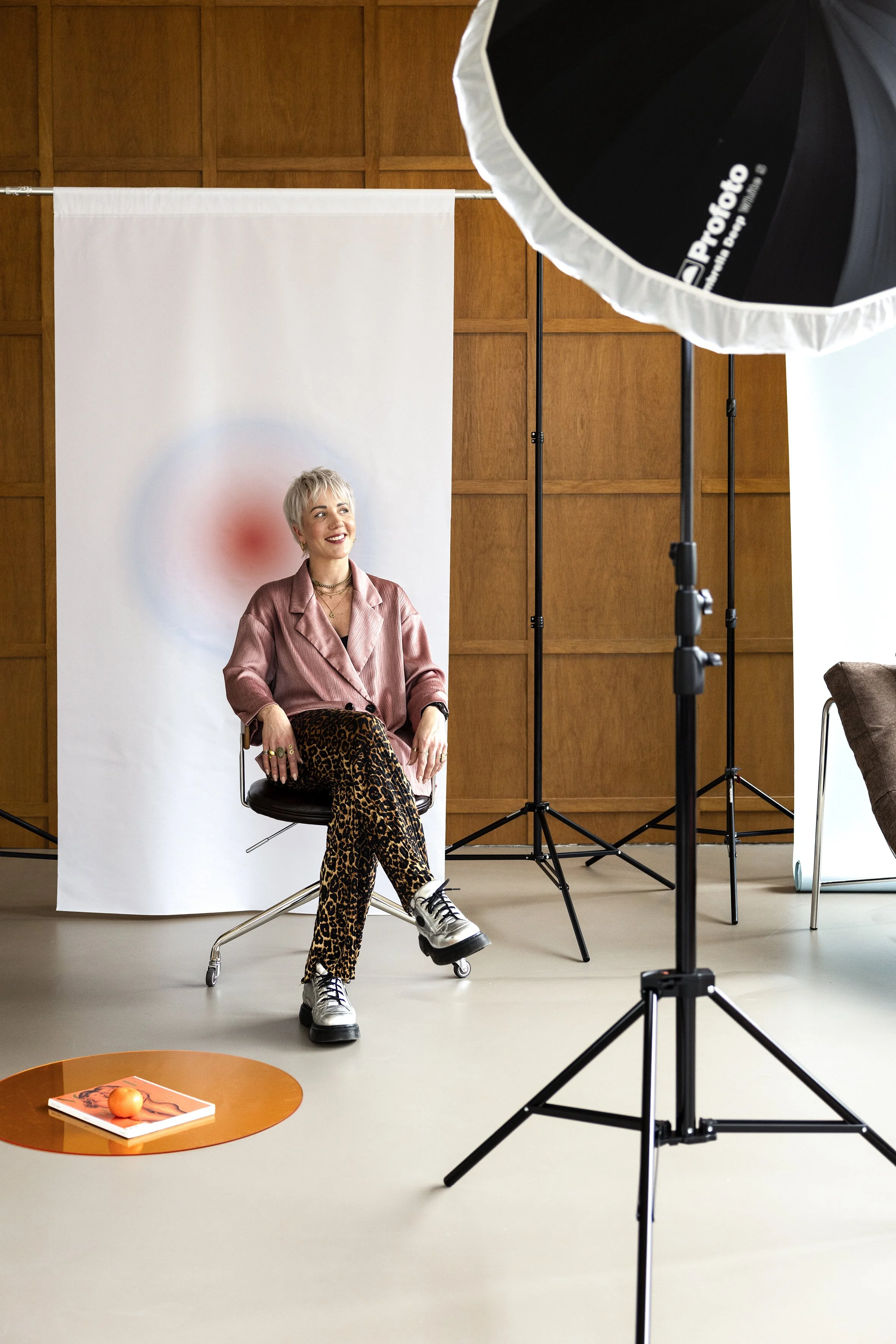 A woman with short blonde hair and a pink satin blazer, wearing leopard print pants and sneakers, sitting on a chair in a photography studio. She is smiling and looking to the left. Behind her is a white backdrop with a red halo effect. The studio ha