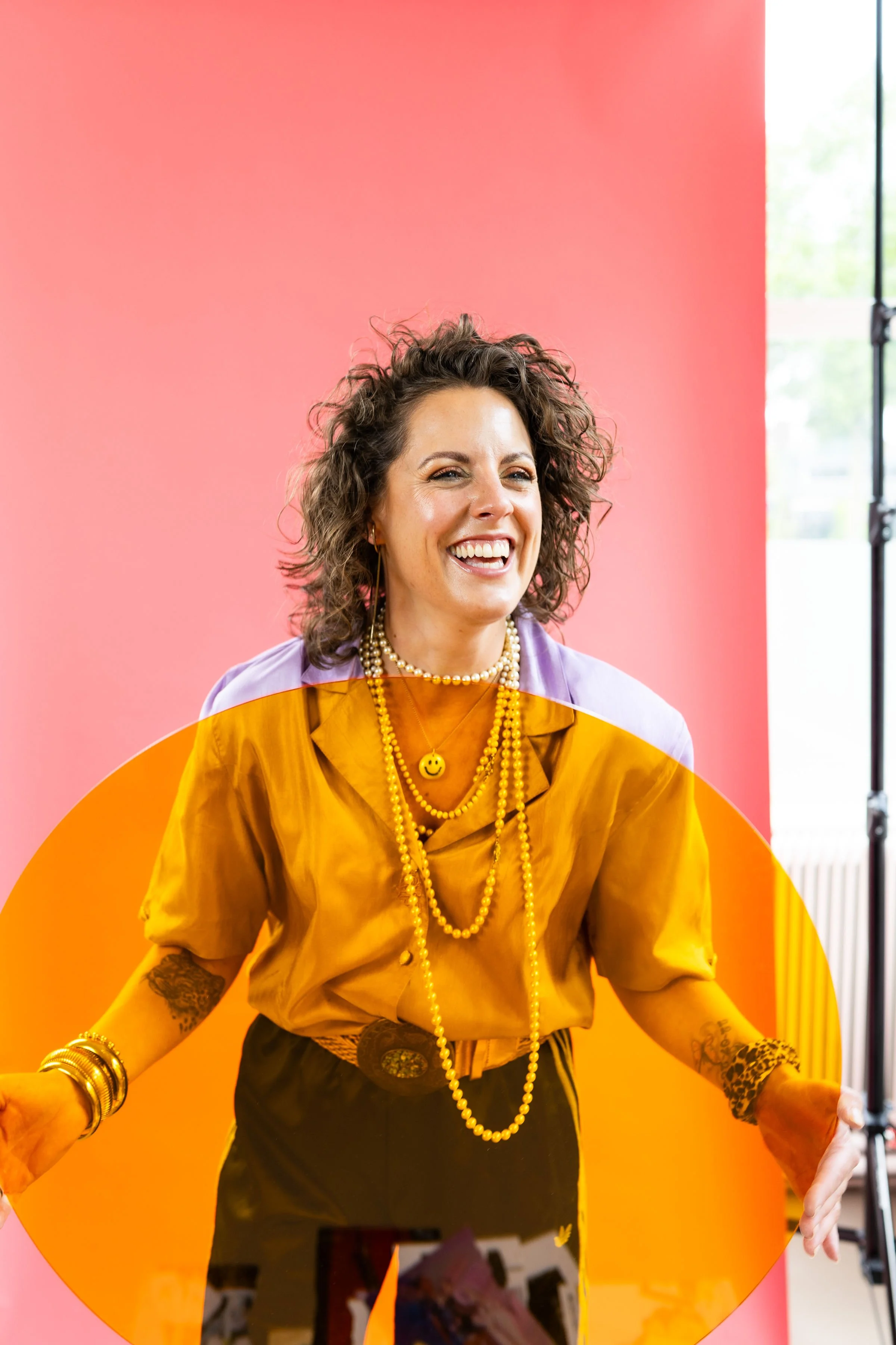 Woman with curly hair wearing a yellow shirt, multiple pearl necklaces, and bracelets, smiling with a pink background and studio lighting.