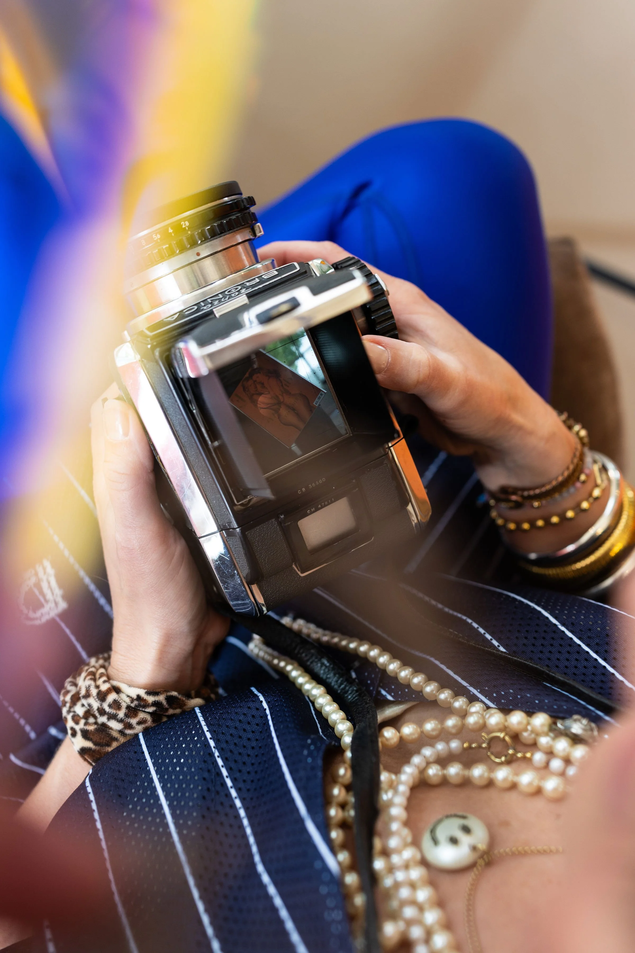 A person holding a camera with a photo of a woman posing on the screen. The person is wearing multiple pearl necklaces, bracelets, and a leopard-print hair tie. The person is dressed in a striped navy and white shirt, and the background includes a blue chair.