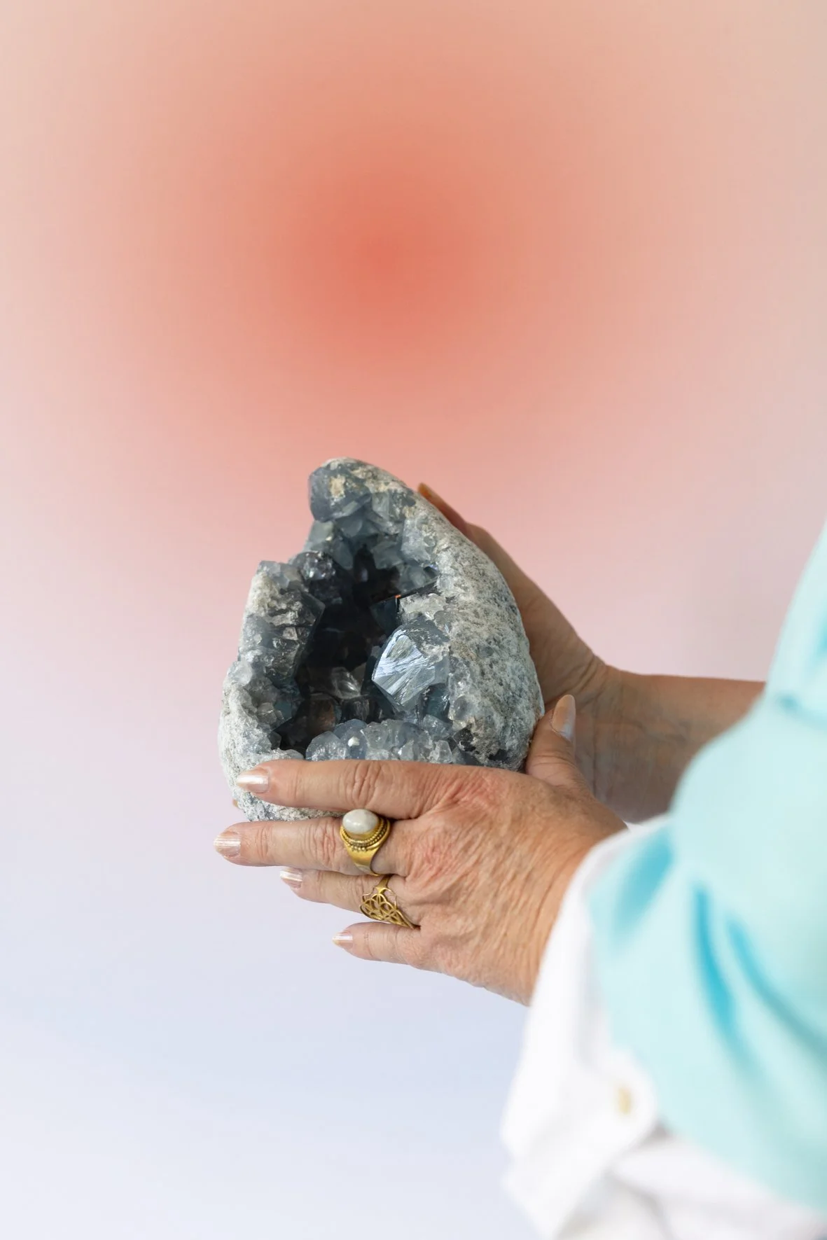 A person holding a gray geode with dark crystals inside, wearing gold jewelry and a light blue sleeve, against a pink gradient background.