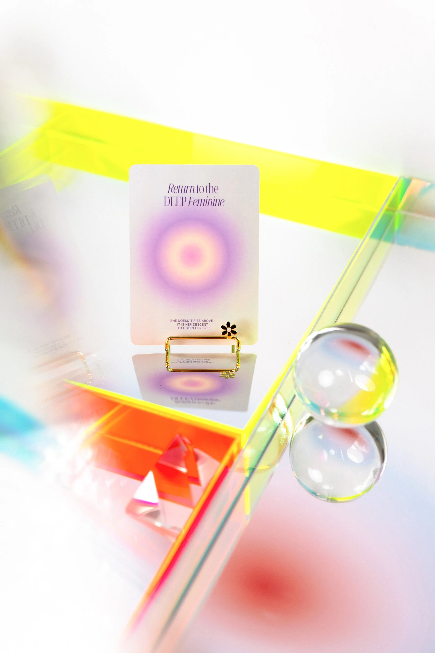Holographic card displays the text "Return to the DEEP Feminine" with a colorful, glowing background, set inside a rainbow-colored geometric display with glass spheres nearby.
