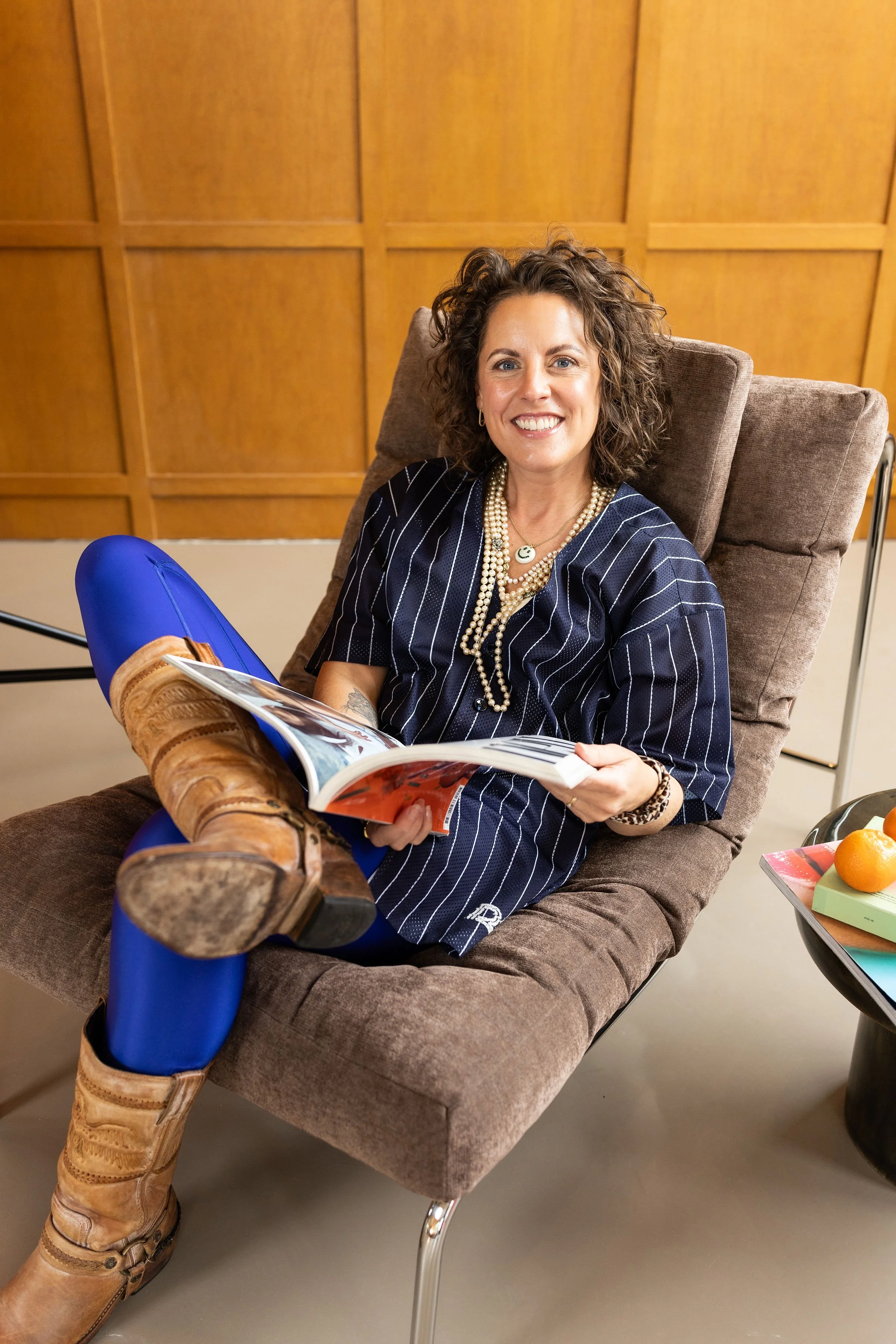 A woman with curly brown hair sitting in a comfortable armchair, smiling, holding a magazine. She wears a navy striped shirt, multiple pearl necklaces, and cowboy boots. The background features a wooden wall, and a small table with oranges and books.