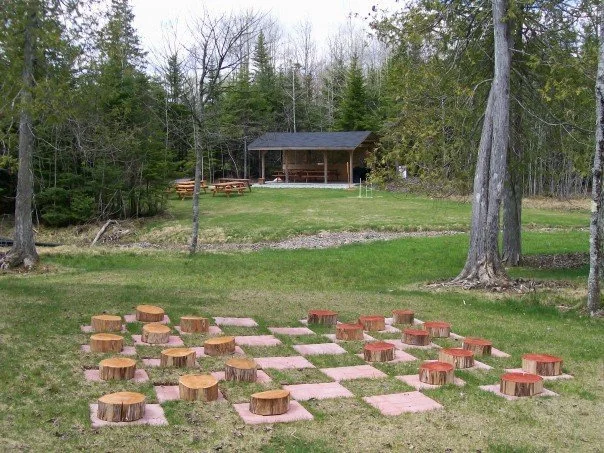 Outdoor scene with a large grassy area, trees, a wooden pavilion in the background, and a giant chessboard with large wooden chess pieces in the foreground.