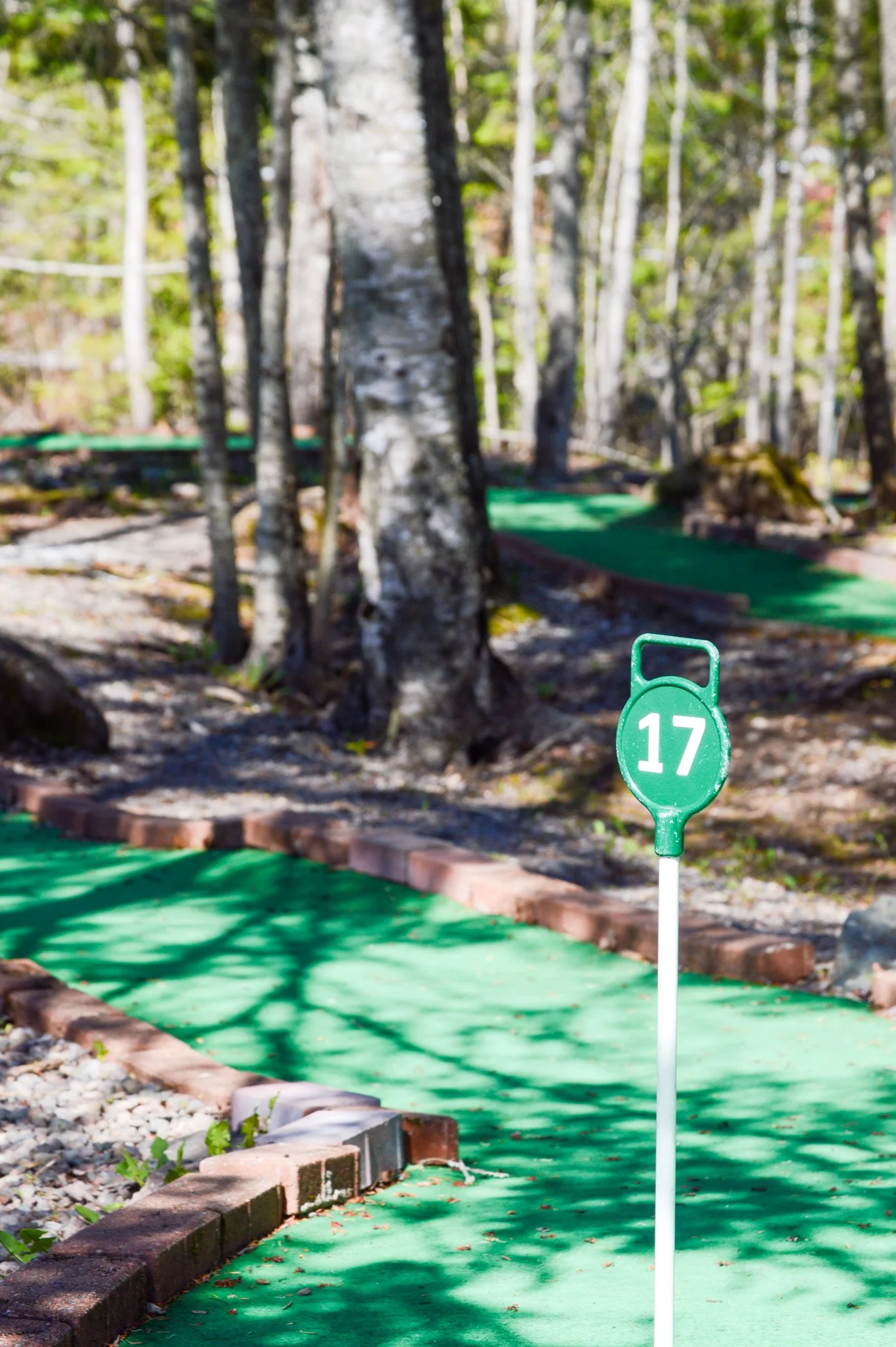 A mini golf course with a green turf, bordered by bricks and small rocks, and a sign showing the number 17. There are trees in the background with sunlight casting shadows.