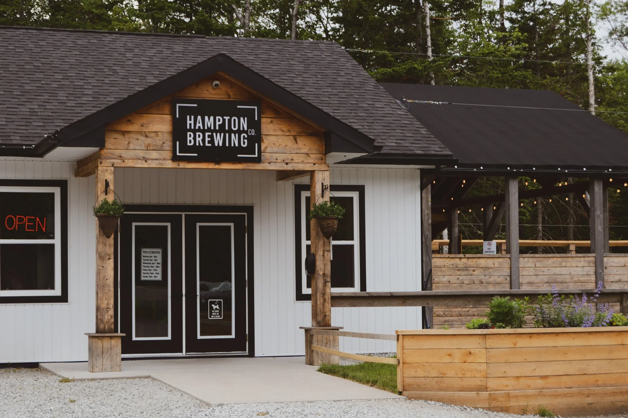 Building with a sign that reads 'Hampton Brewing Co.' in a rural setting with trees in the background. The building has a gray shingled roof and a black and white facade with a small patio area featuring hanging plants and string lights.