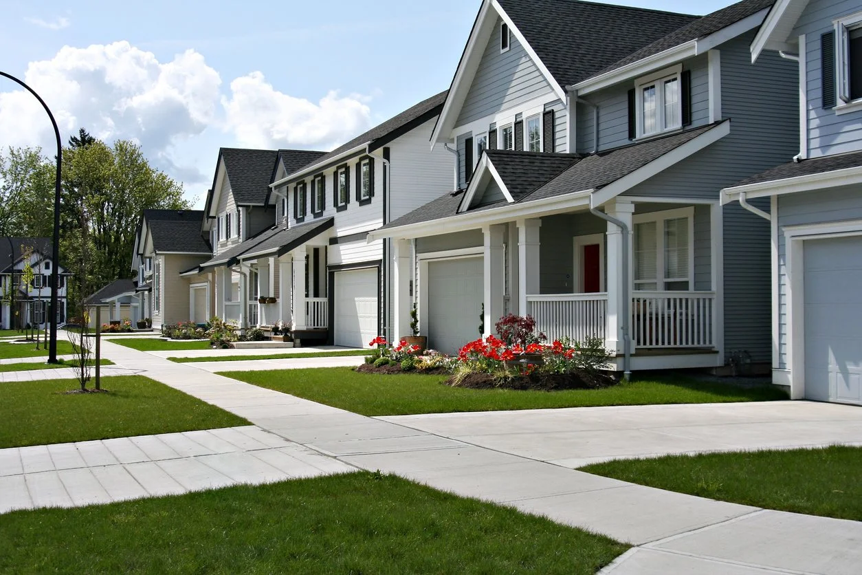 Series of modern suburban houses with well-maintained lawns and flower beds, sidewalks, and a clear sky in the background.