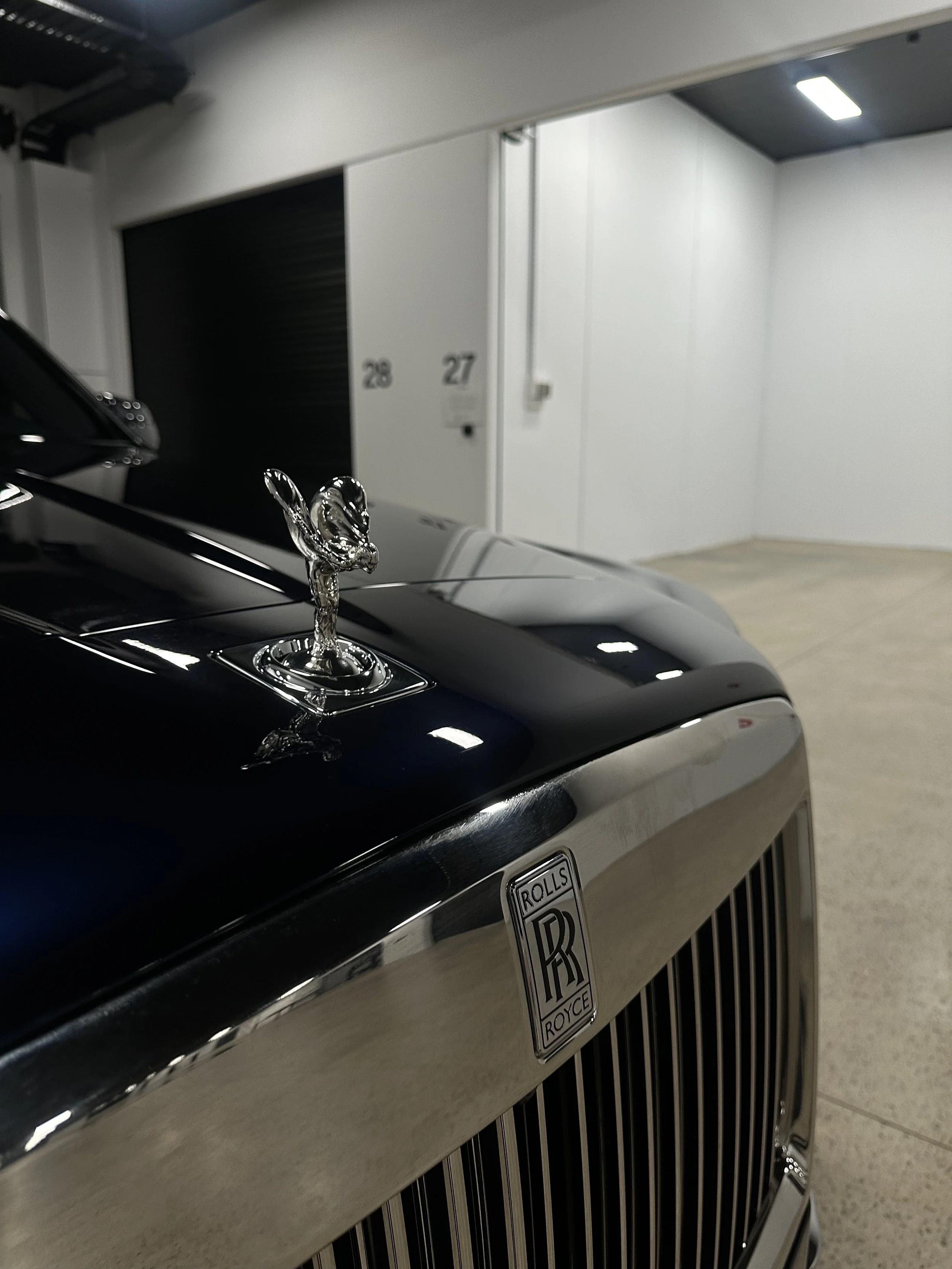 Close-up of a black Rolls-Royce car with a silver hood ornament, parked in an indoor garage with white walls and numbered doors.