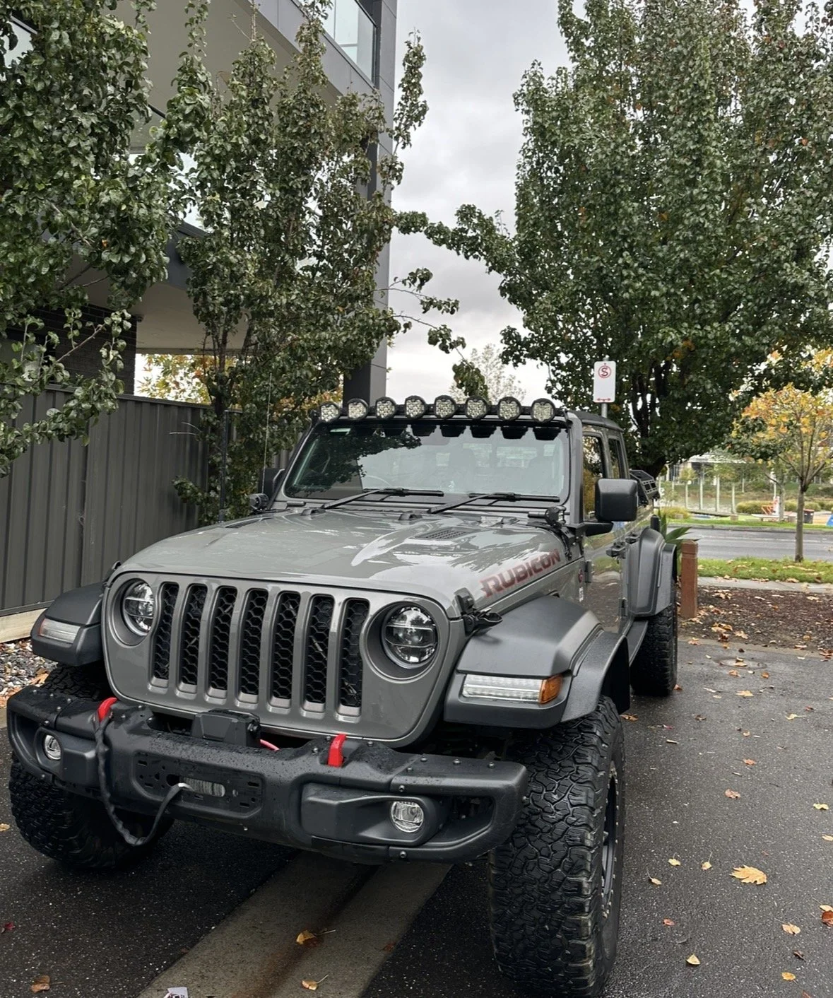 Gray Jeep Rubicon parked in a lot with trees and a modern building in the background, overcast sky, and fallen leaves on the ground.