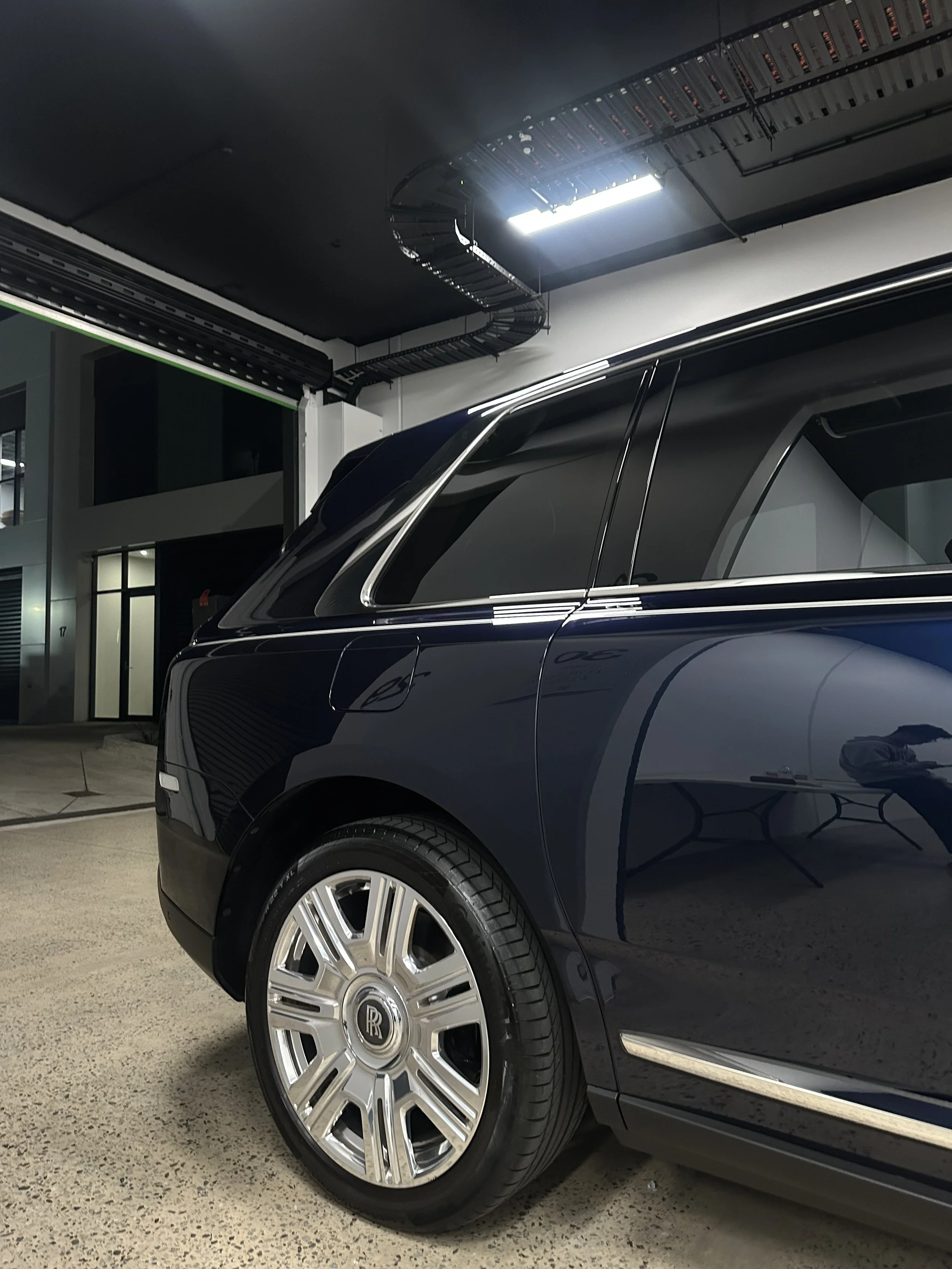 Close-up of a black luxury car (Rolls Royce) parked indoors, showing the front wheel and part of the body, with a building and lighting visible in the background.