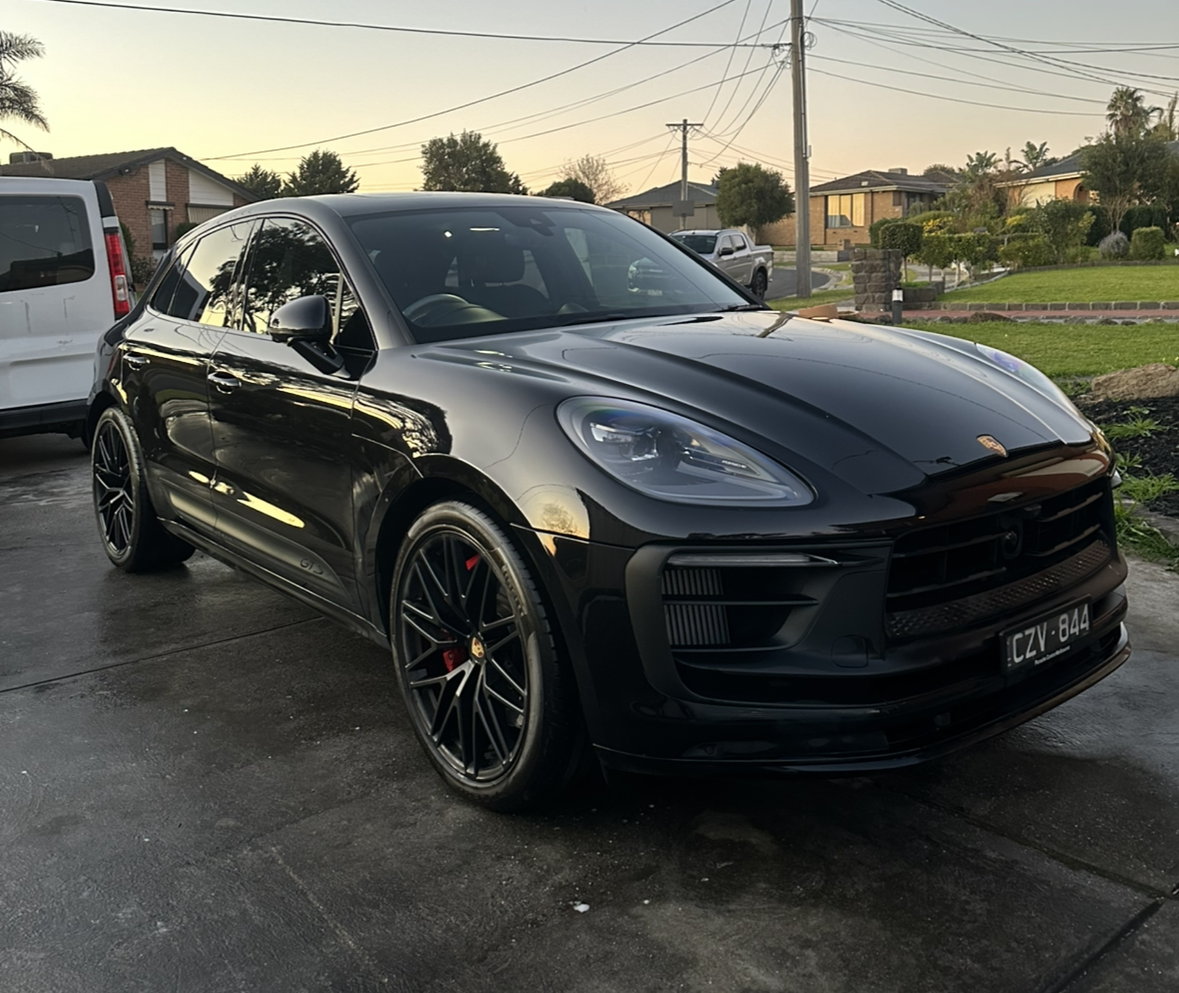 Black Porsche SUV parked on a driveway with neighboring houses and a sunset sky in the background.