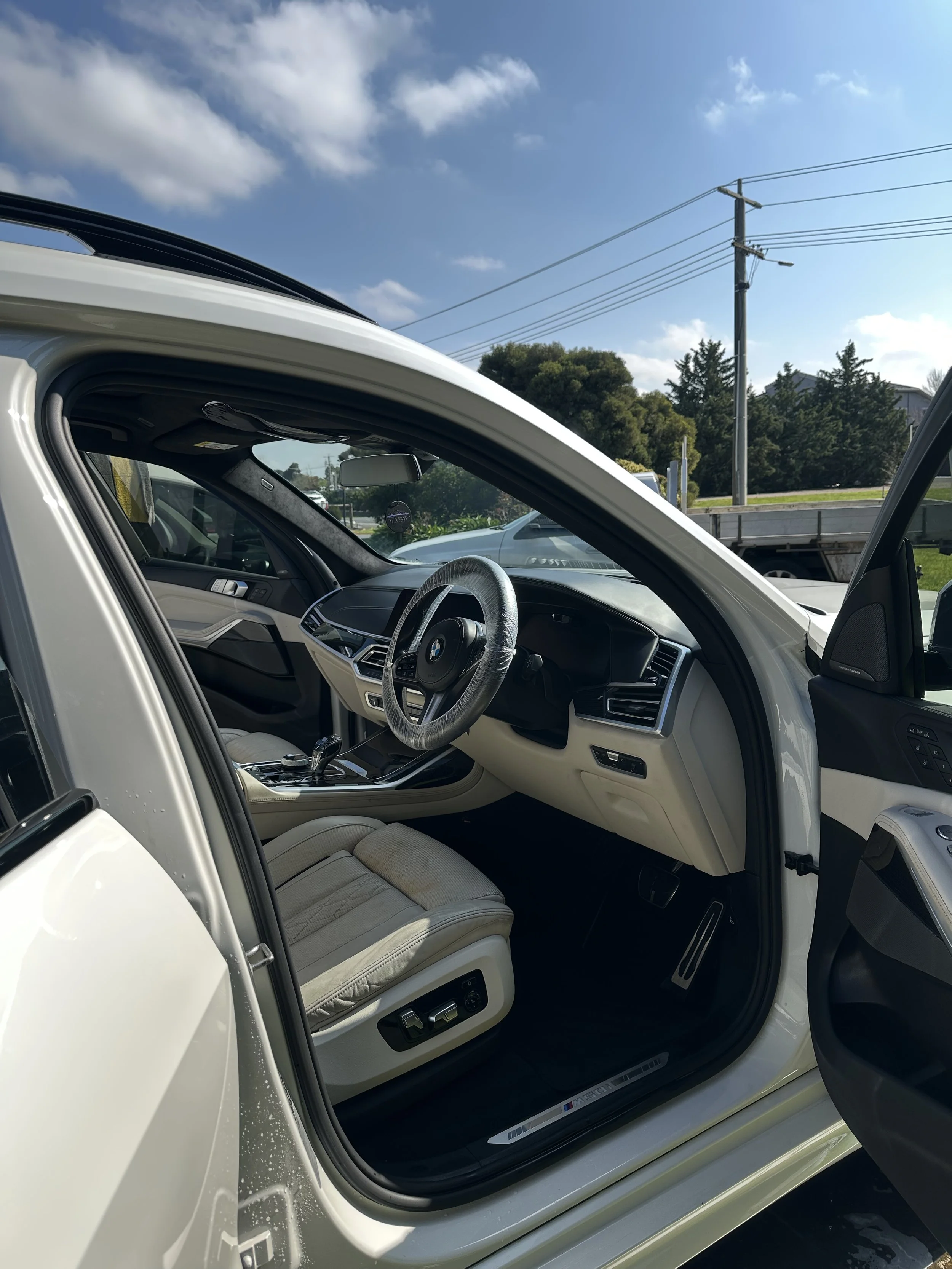 Interior of a white luxury BMW vehicle with a beige leather seat, steering wheel, and dashboard, seen from an open driver's side door. Outside, a parking lot with other vehicles, a utility pole, trees, and a partly cloudy sky are visible.