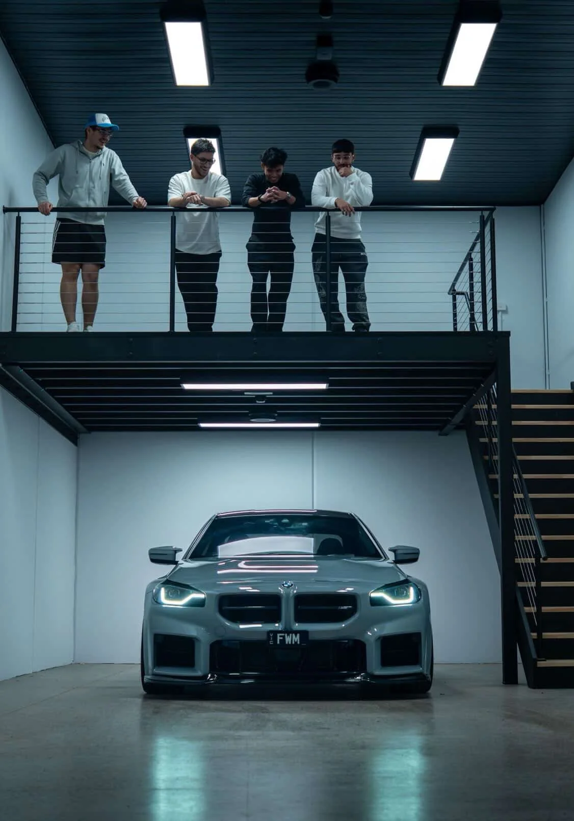 Four young men standing on a mezzanine with a black railing, looking down at a silver sports car parked in a garage below. The garage has plain white walls, a concrete floor, and black overhead lighting.