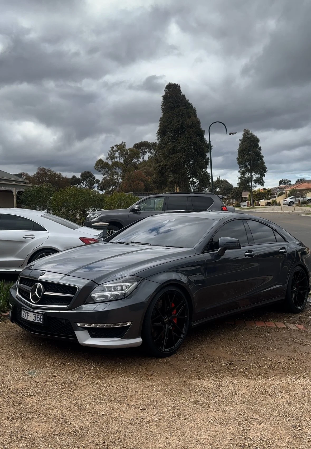 A parking lot with several cars, including a black Mercedes-Benz with tinted windows in the foreground, under a cloudy sky with tall trees and houses in the background.