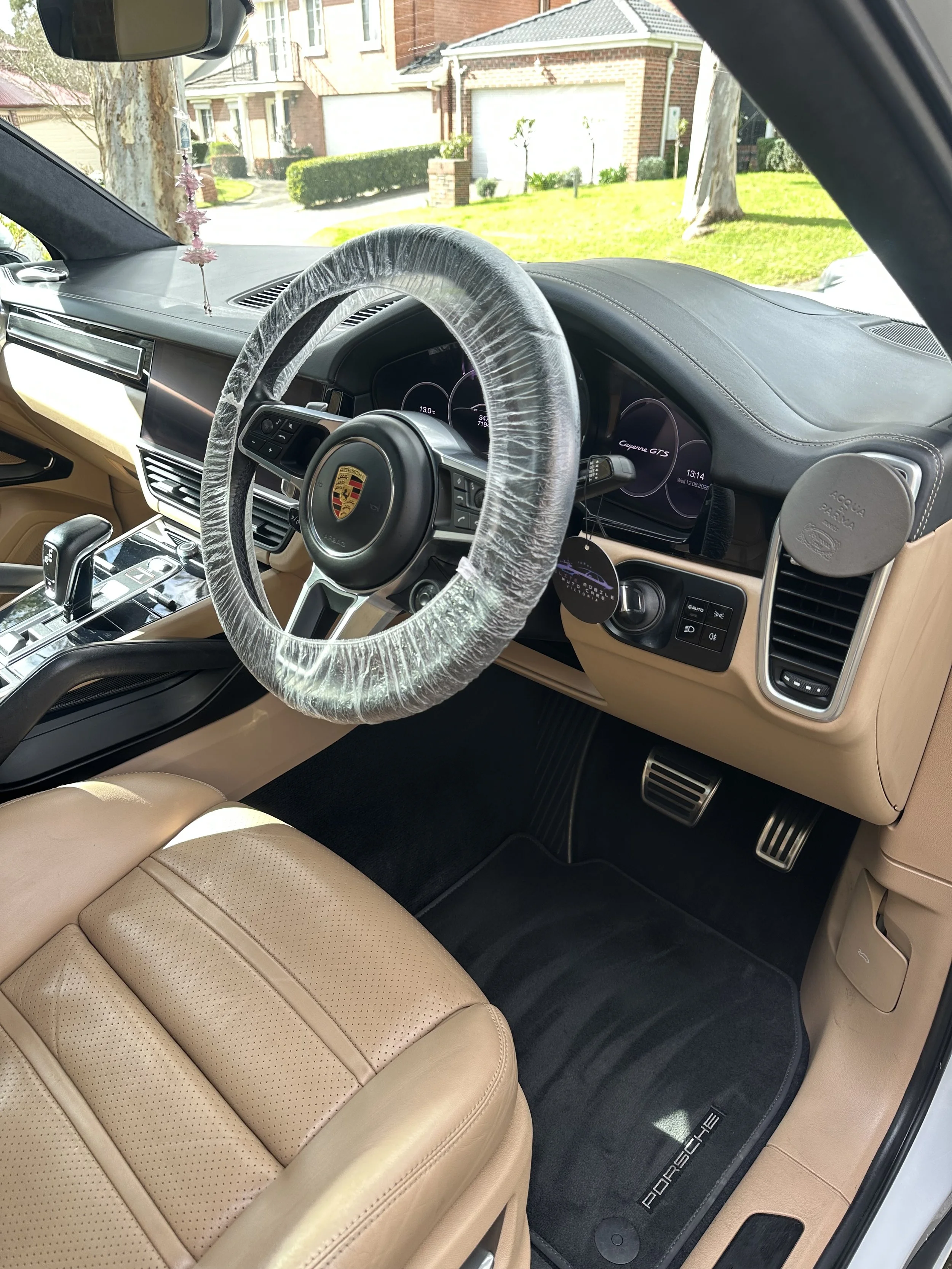 Interior of a luxury car, showing a beige leather seat, a black floor mat with the Porsche logo, a steering wheel wrapped in plastic, and digital dashboards displaying the car model as Porsche Cayenne GTS.