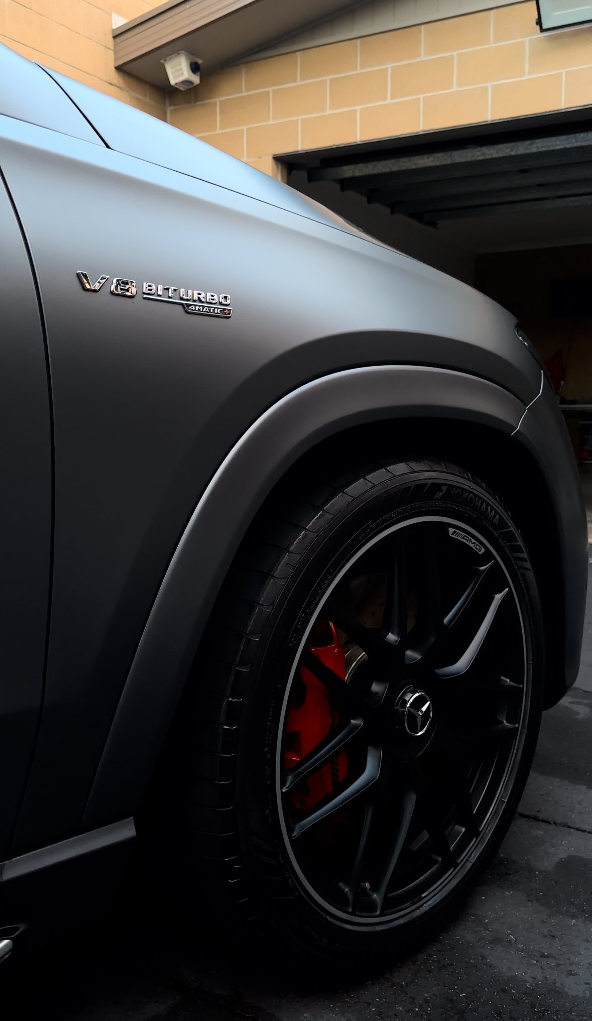 Close-up of a gray Mercedes-Benz car with AMG wheels, a red brake caliper, and a badge indicating V8 Biturbo 4MATIC engine, parked in a garage with brick wall and ceiling camera in the background.