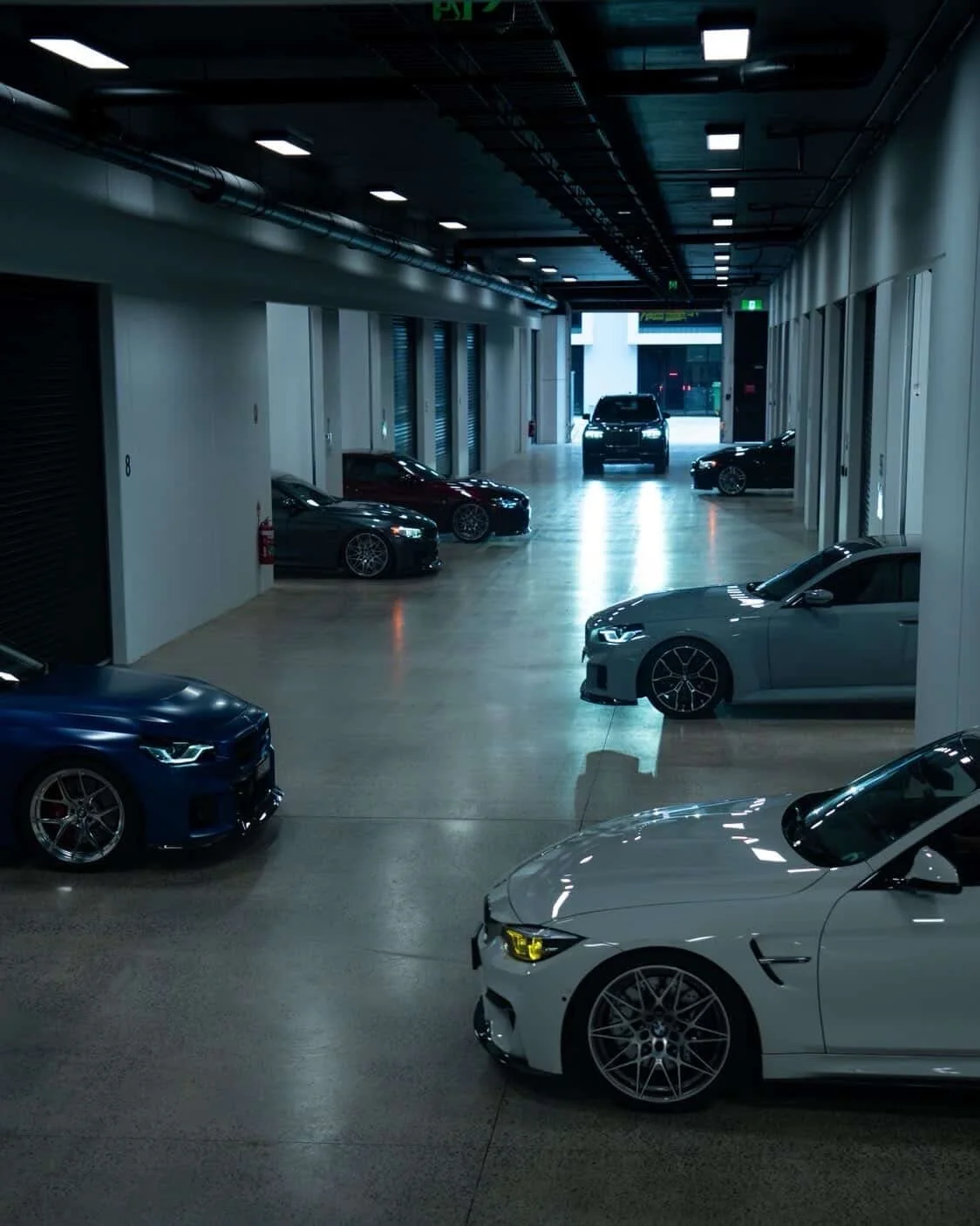 Indoor parking garage with several luxury cars parked along the walls and in the open space, illuminated by ceiling lights with an exit visible in the distance.
