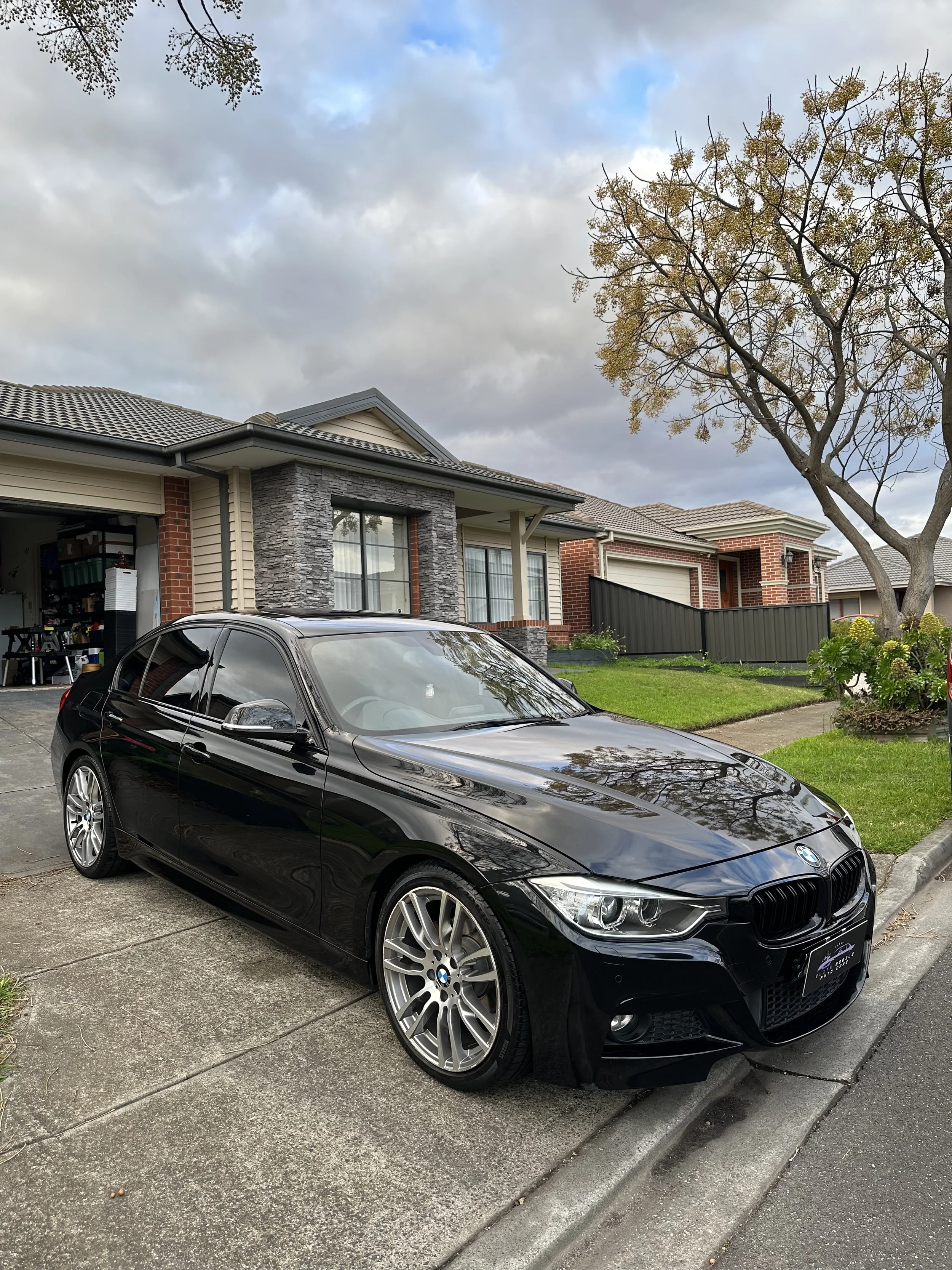 A black BMW car parked on the driveway in front of a residential house, with a garage open showing various tools and items inside. The house has a brick and beige siding exterior, with a stone pillar near the window, and a tree with sparse leaves in the yard. The sky is cloudy.