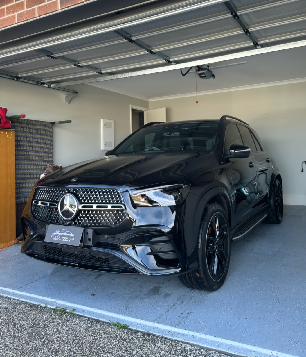A black Mercedes-Benz SUV parked inside a residential garage with a partly open garage door, a dresser and a wrapped item in the corner.