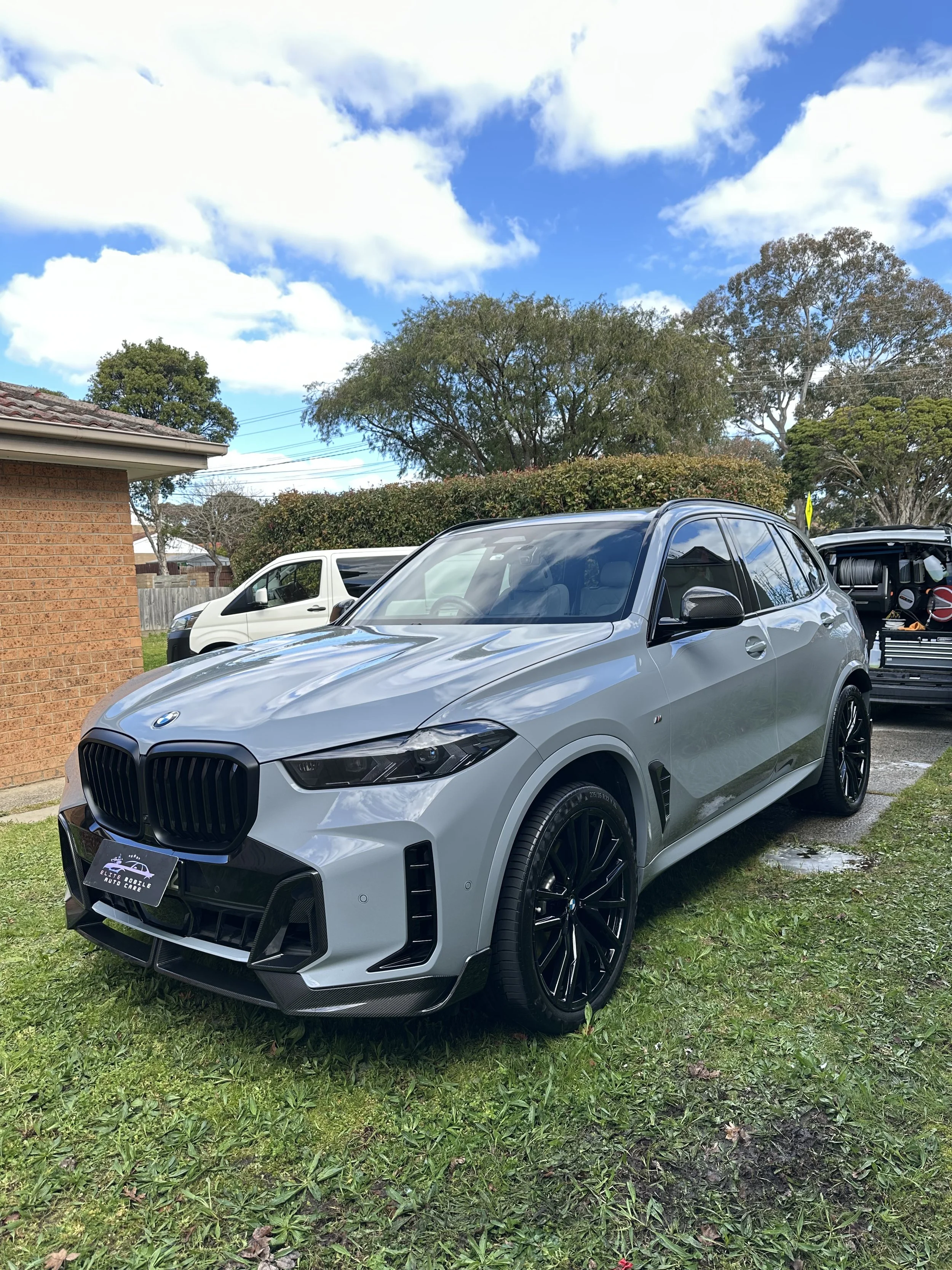 A silver BMW SUV parked on a grassy area in front of a house, with trees and blue sky with clouds in the background.